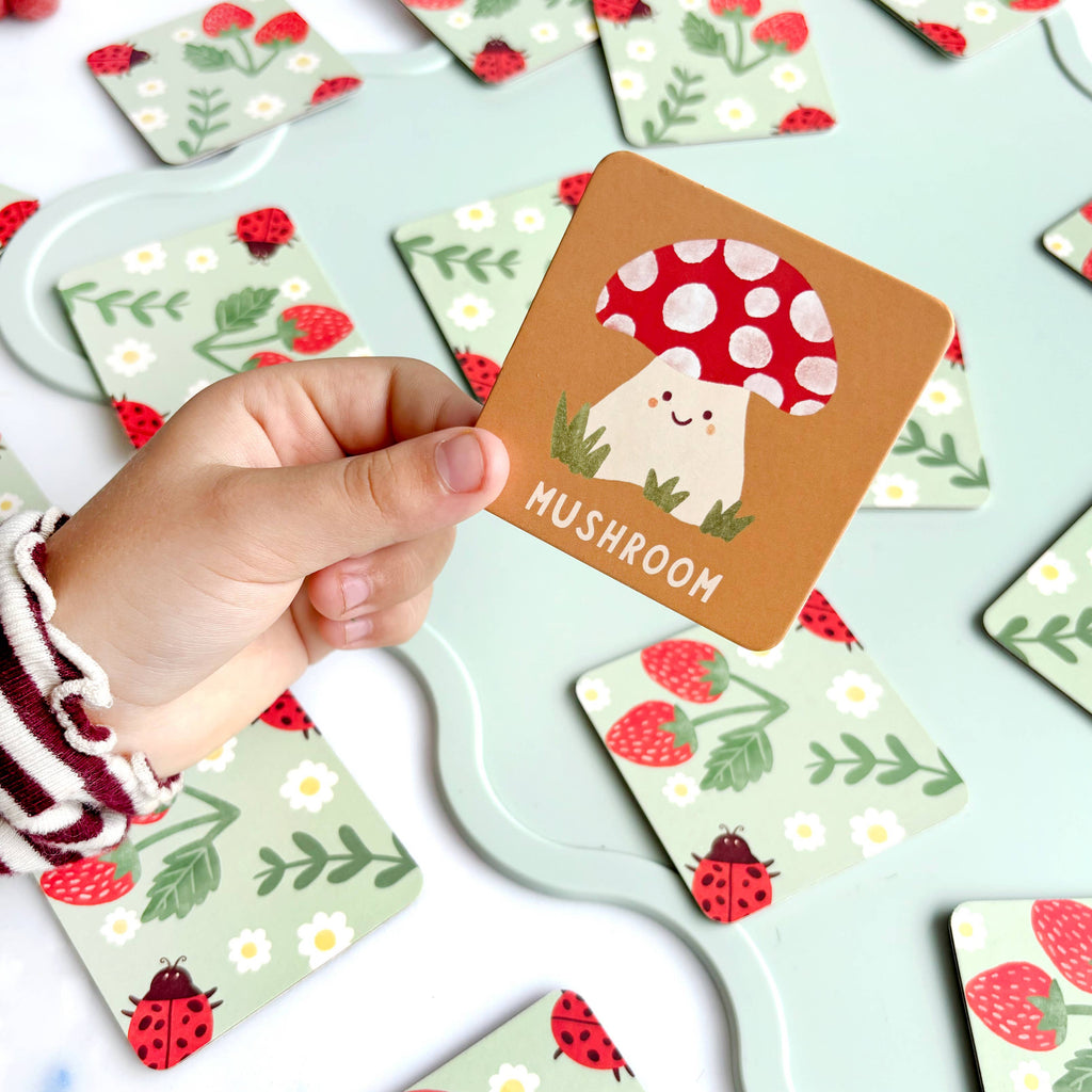Hand holding a card with a mushroom design among other nature-themed cards on a light blue surface.