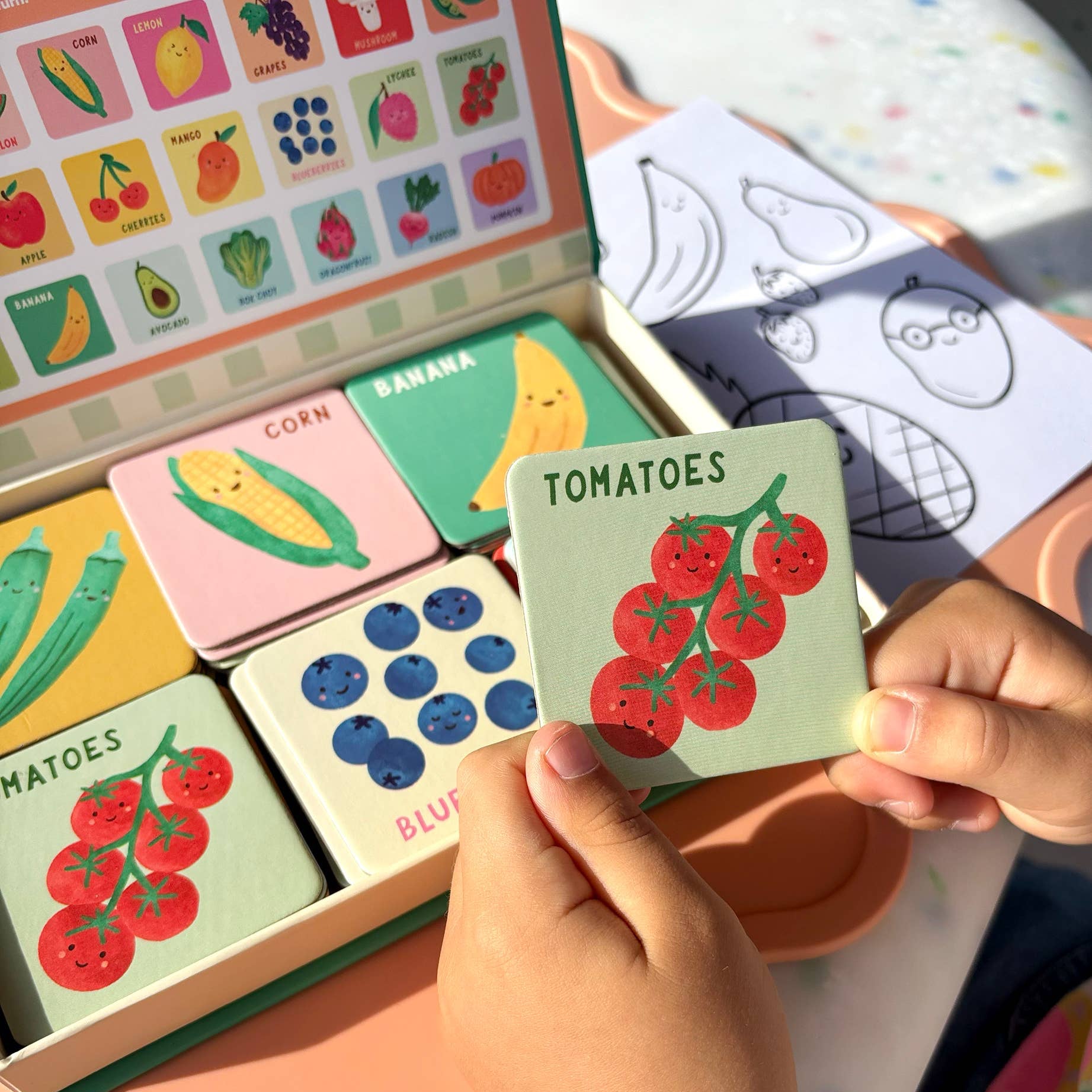 Child holding a card with 'Tomatoes' next to a set of educational cards with illustrations of fruits and vegetables.