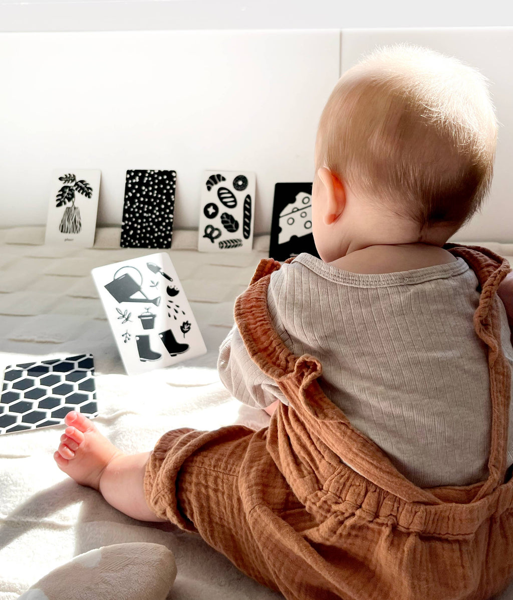 Baby wearing brown overalls sitting on a light-colored surface with framed black and white art prints in the background.