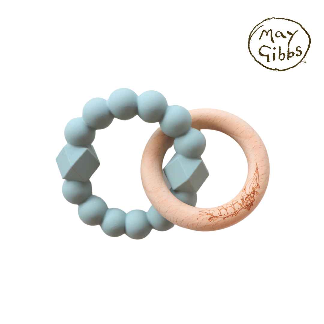 A sage-colored silicone teething ring attached to a beech wood teething ring, both designed by May Gibbs.