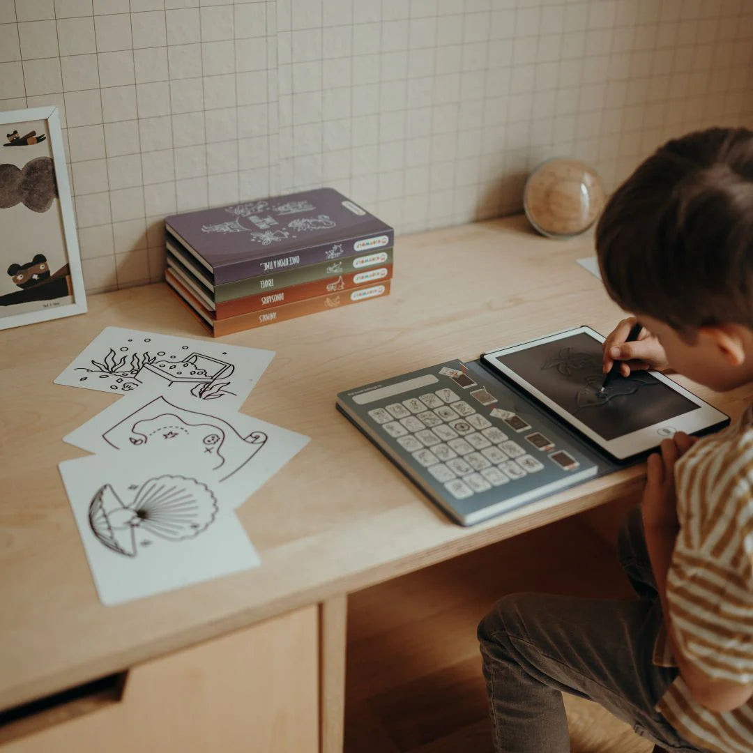 Child using a tablet with drawing paper and books on a wooden table.
