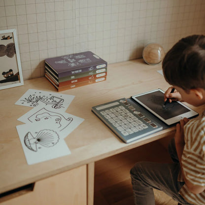 Child using a tablet with drawing paper and books on a wooden table.