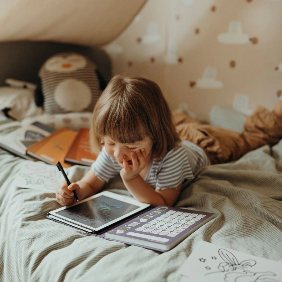Child lying on a bed with books and a tablet, surrounded by cozy bedding and pillows.