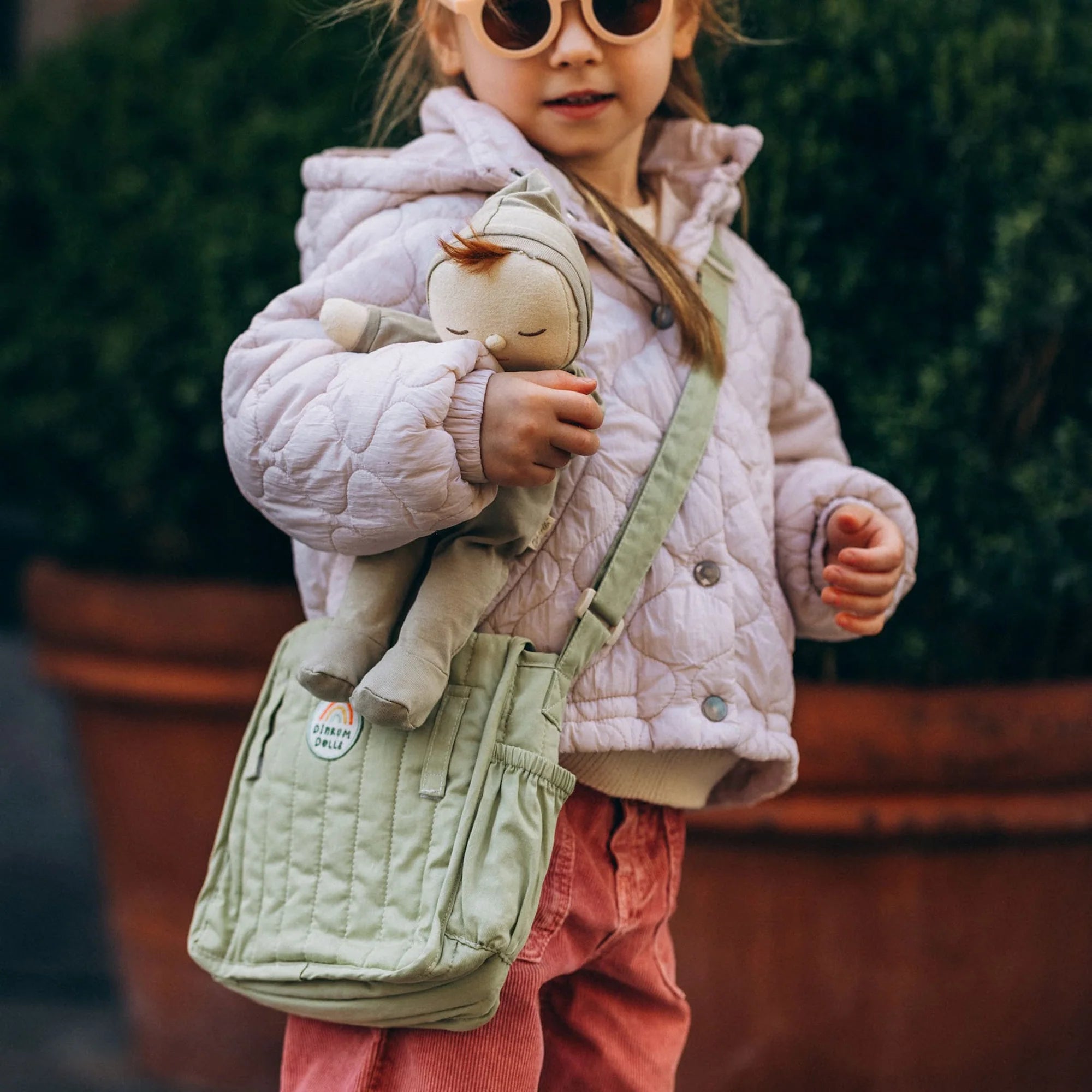 Child holding a plush toy and a green bag with a plant in the background