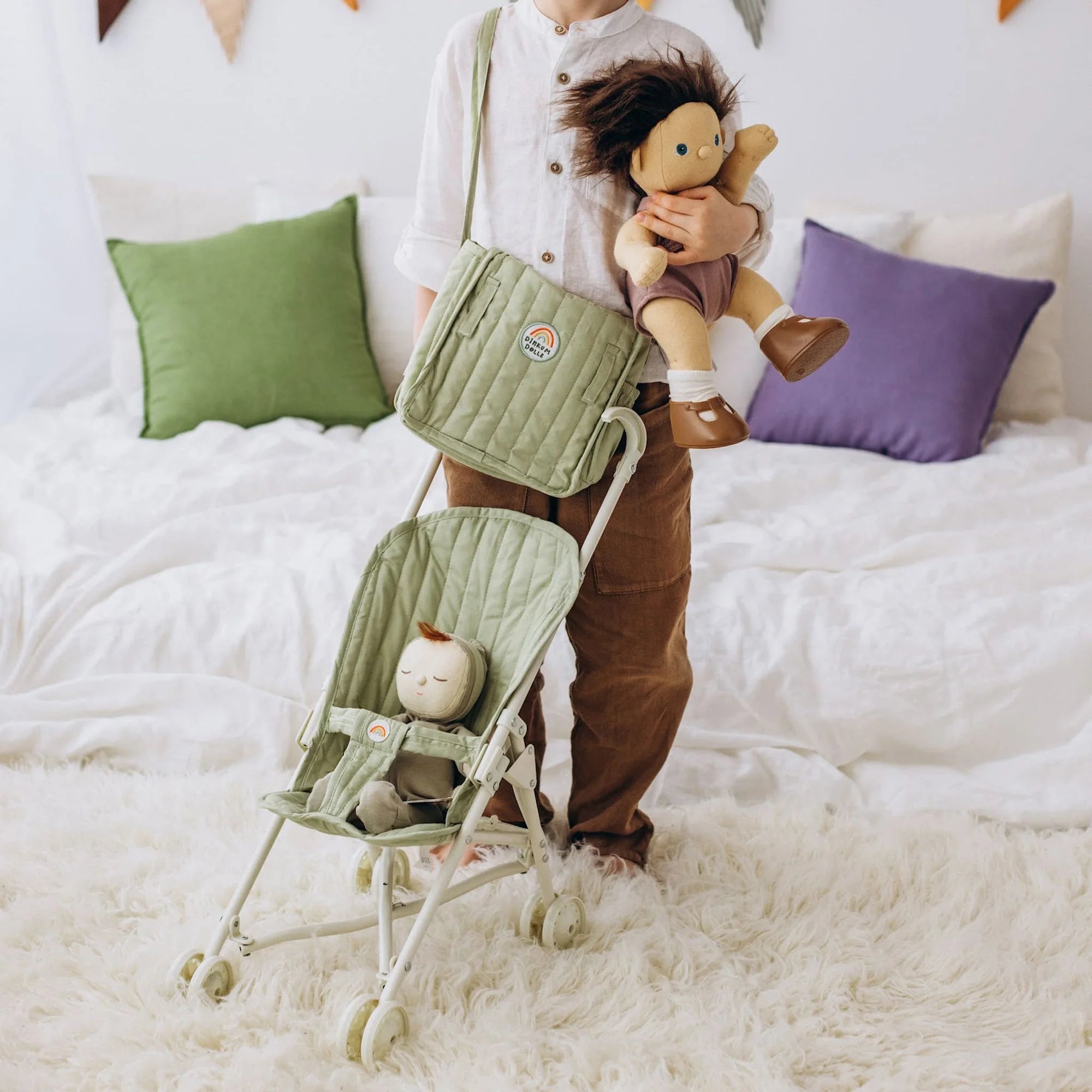 Person holding a babydoll next to a small green stroller on a white bed with colorful pillows.