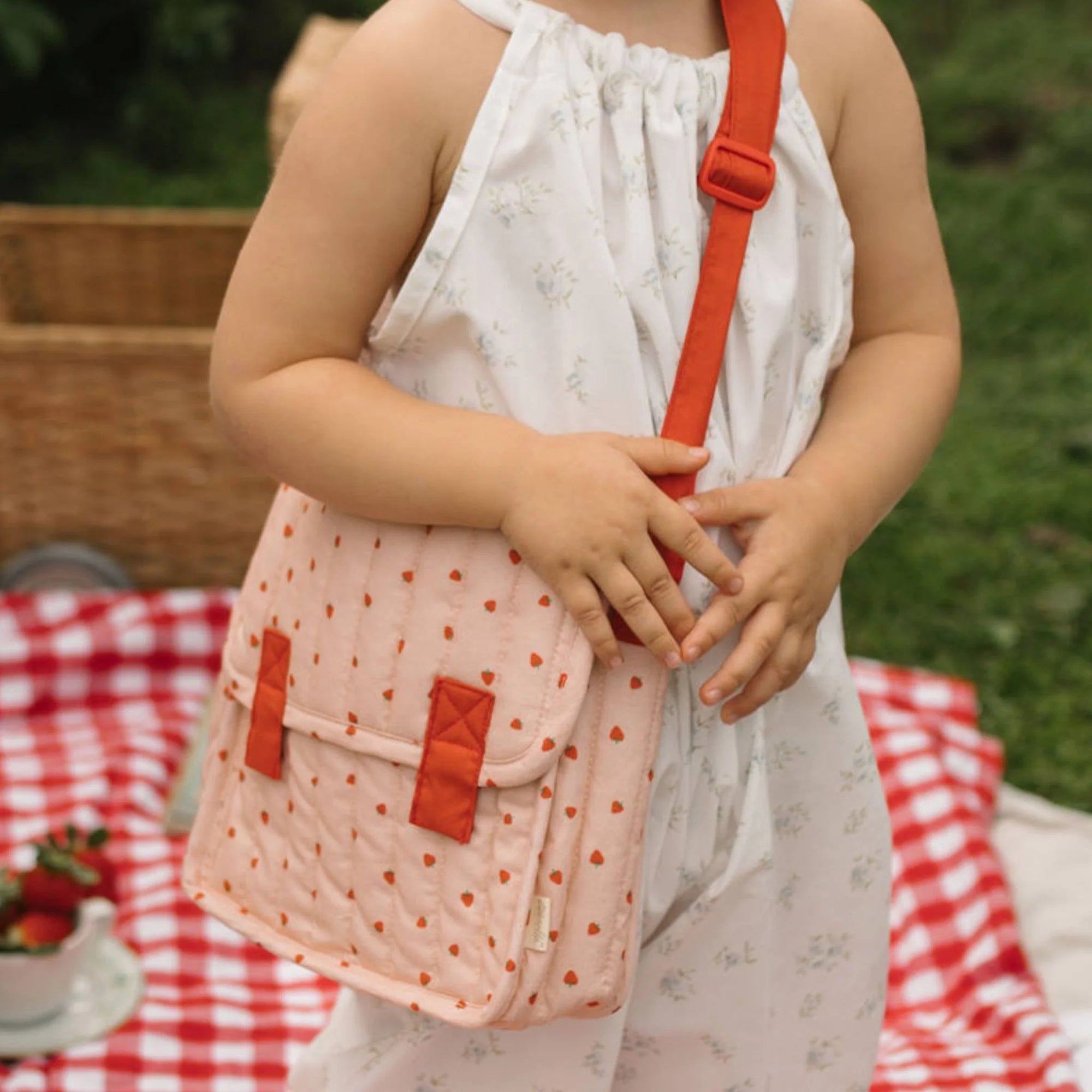 Child holding a pink polka dot bag with red straps on a picnic blanket