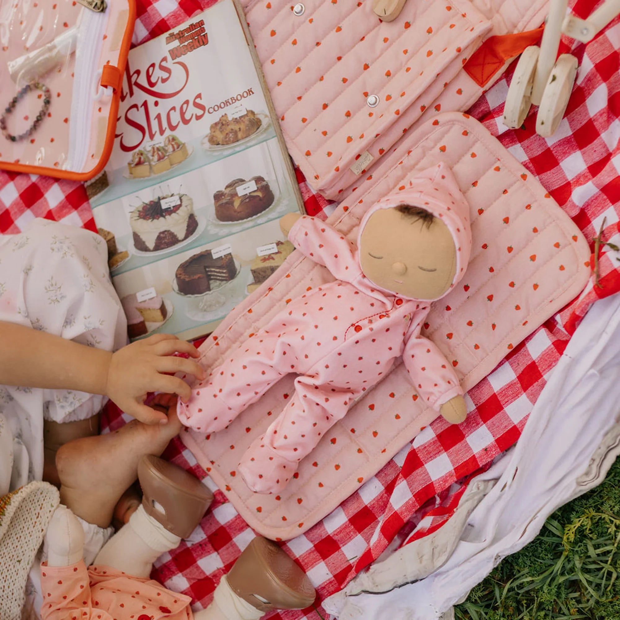 Child playing with a doll on a red and white checkered blanket, surrounded by a cookbook and other toys.