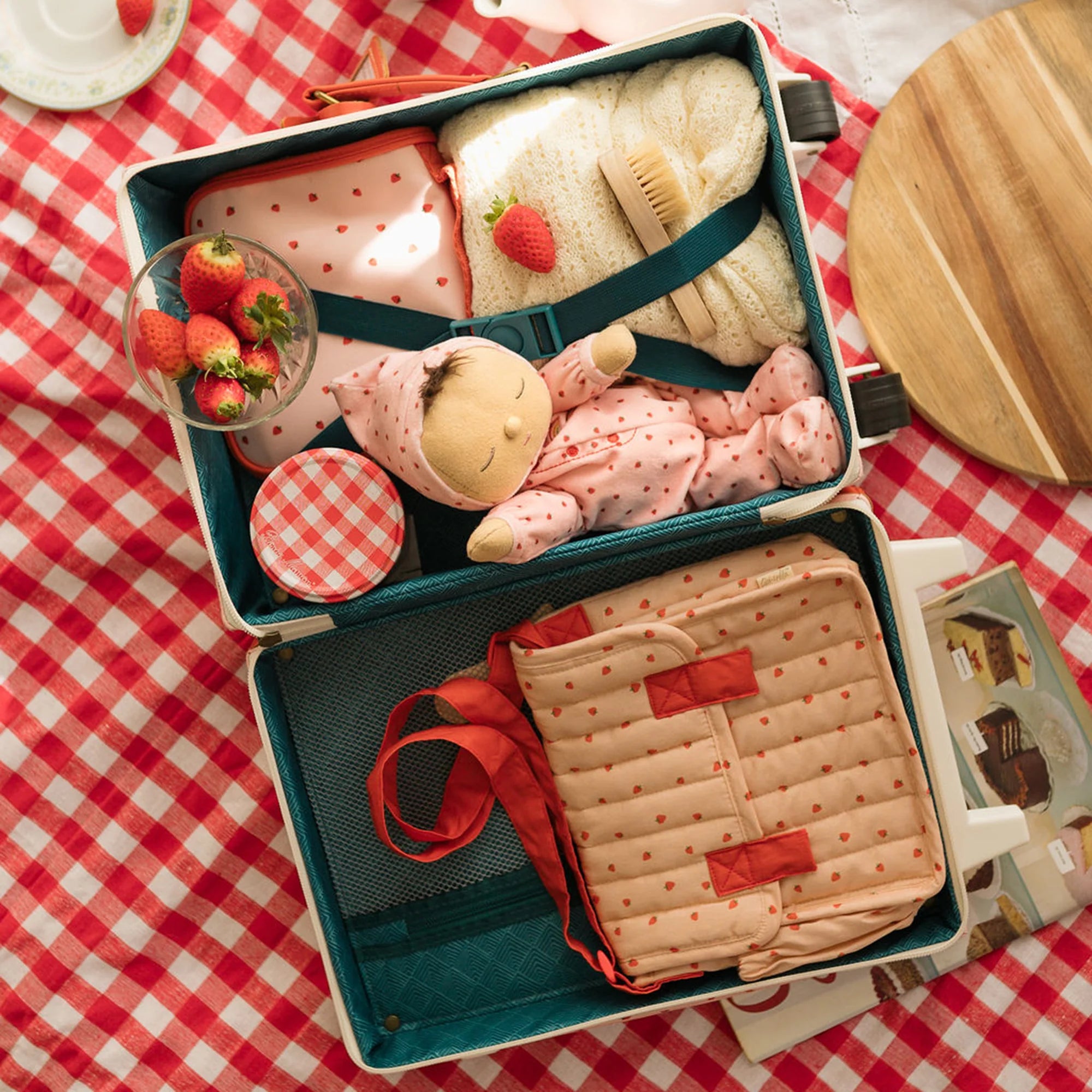 Toy suitcase with teddy bear, strawberries, and other items on a red and white checkered tablecloth.