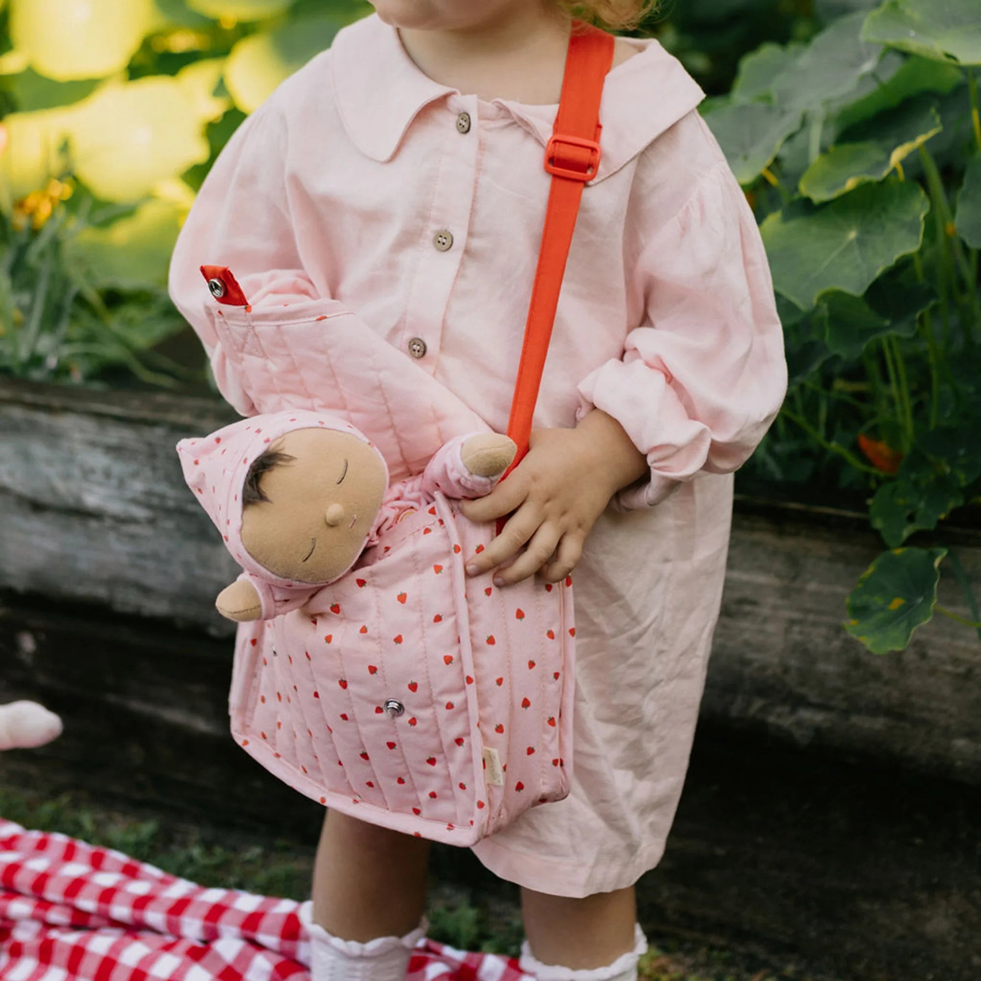 Child holding a pink backpack with a doll, standing outdoors.