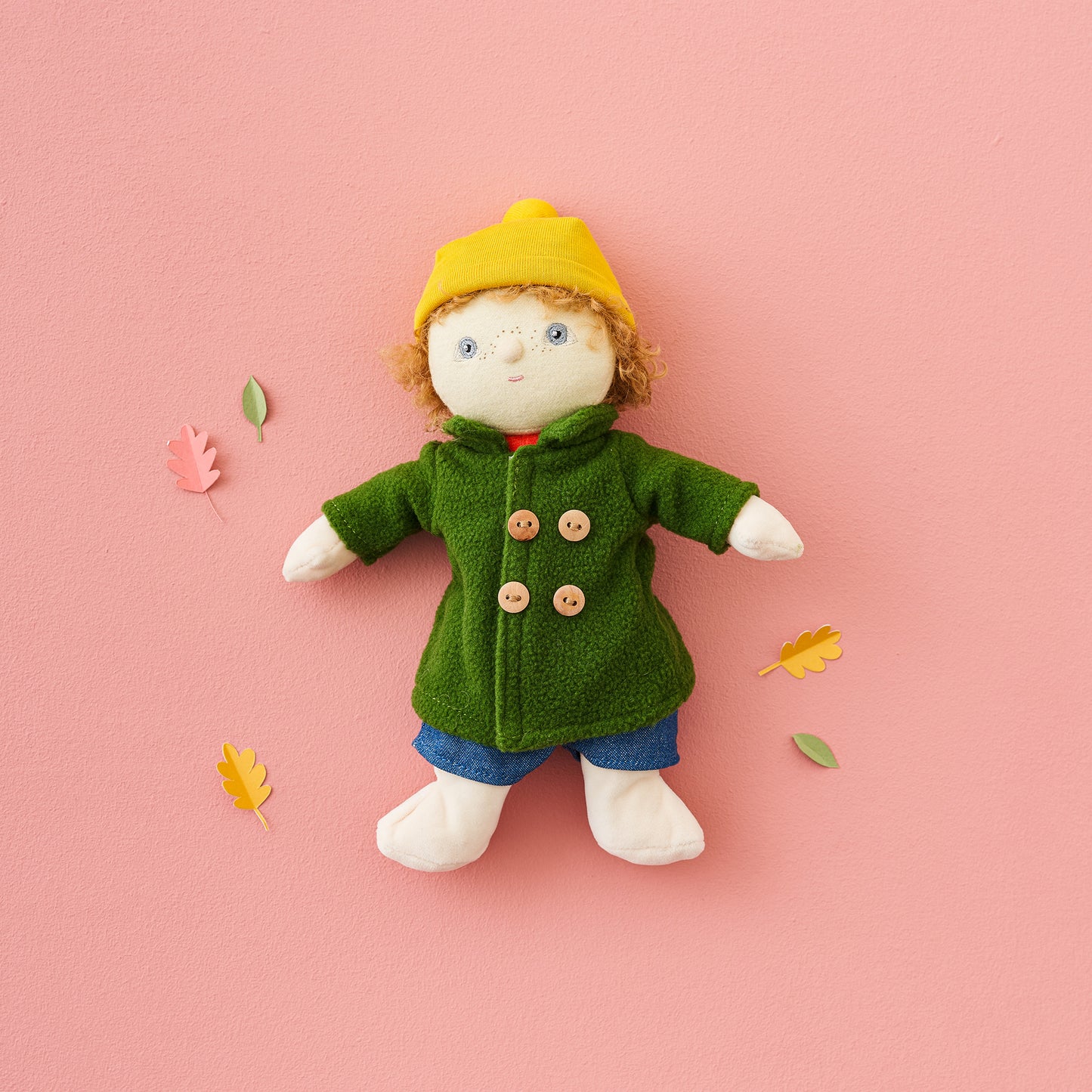 Toy figure wearing a green coat and yellow hat on a pink background with scattered leaves.