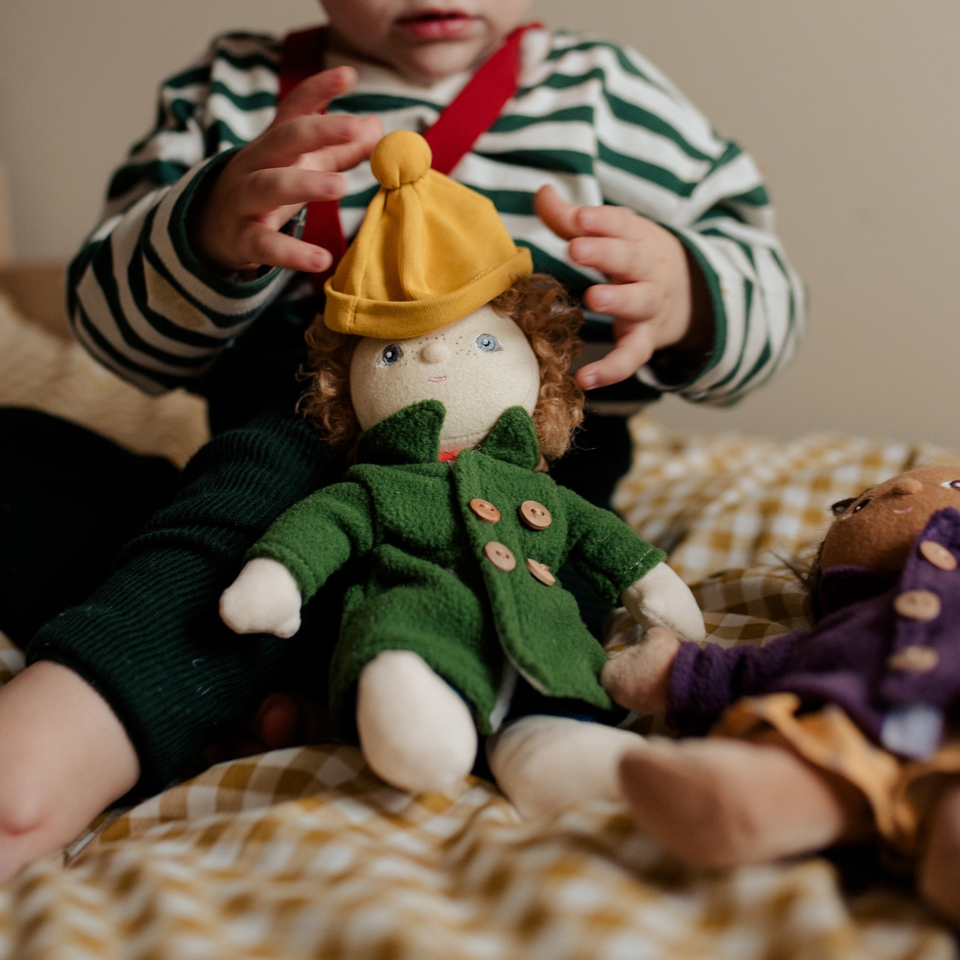 Child holding a doll wearing a yellow hat and green coat on a checkered blanket