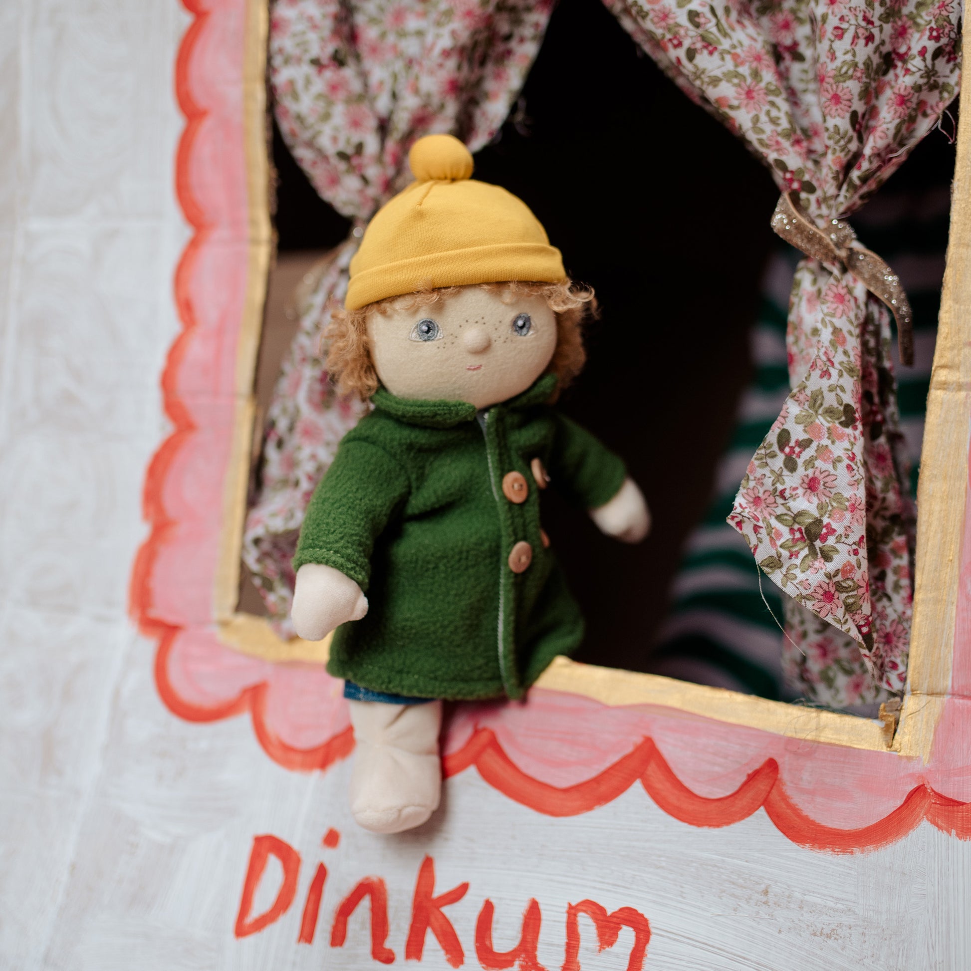 Small doll in a green coat and yellow hat standing inside a wooden frame with floral curtains.