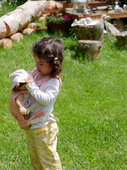 Child holding a doll outdoors on grass with logs and plants in the background