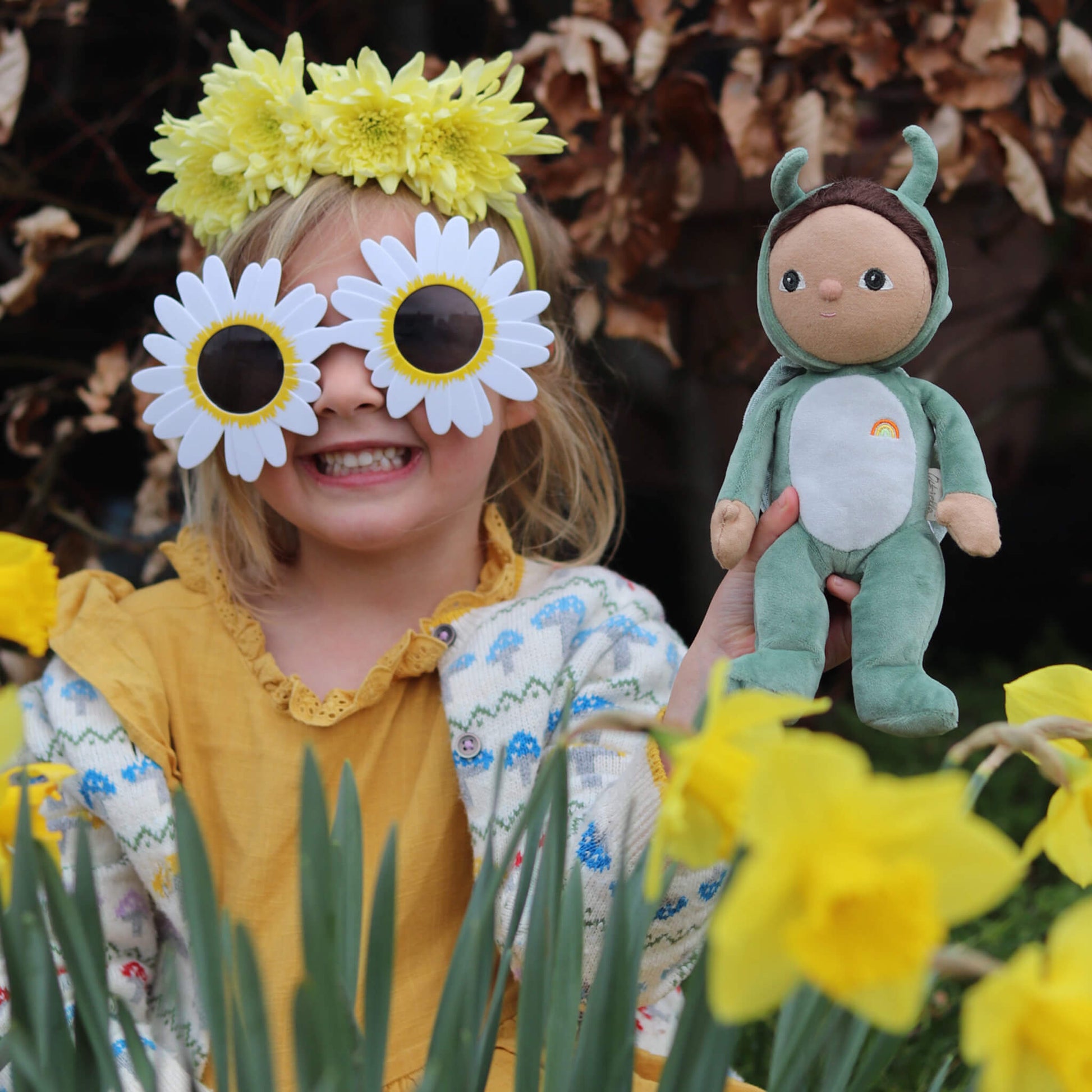 Child wearing flower headband and sunglasses holding a doll amidst yellow flowers