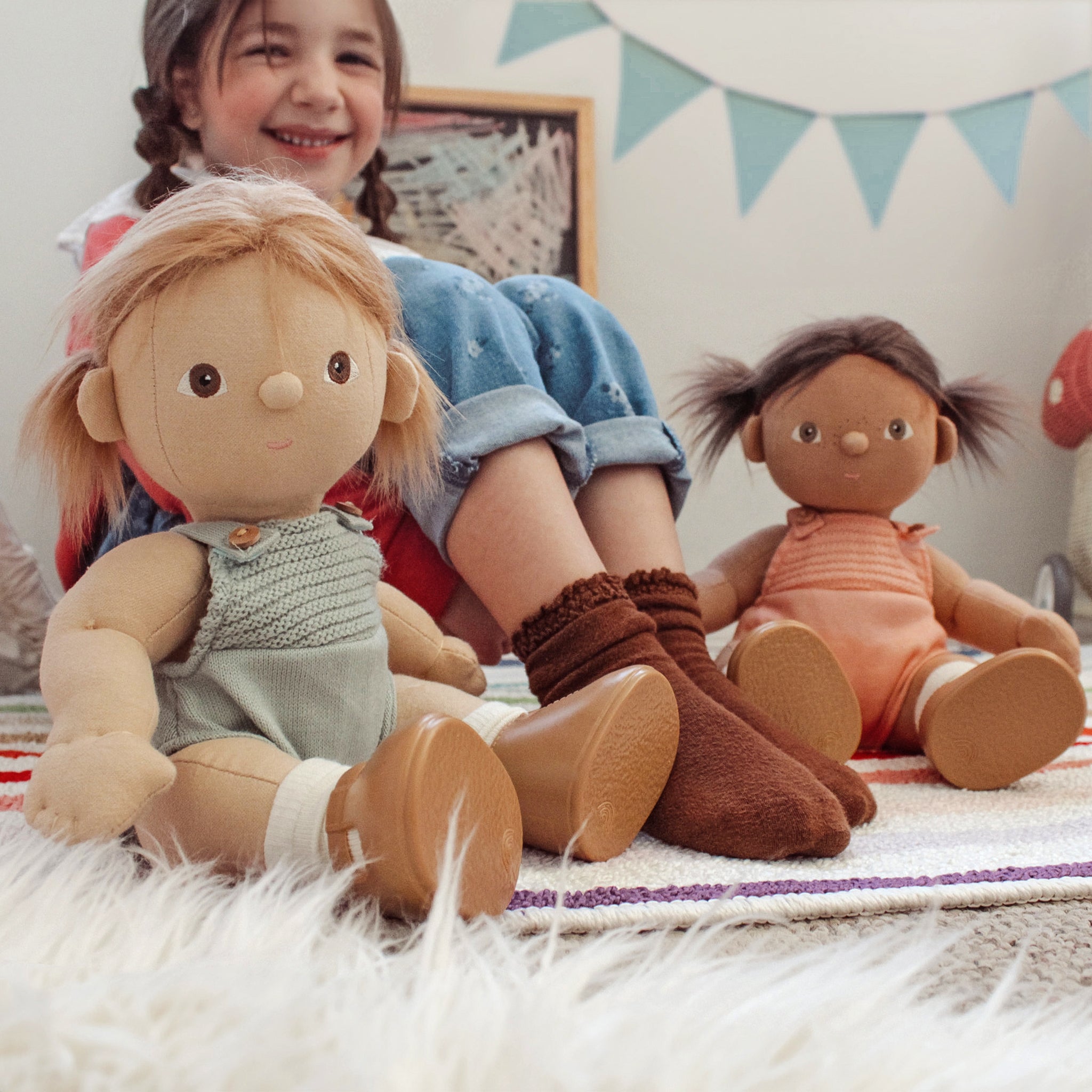 Child sitting with two dolls on a carpeted floor, surrounded by decorative elements.