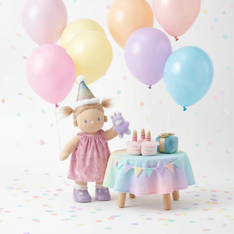 Doll with a birthday hat surrounded by colorful balloons and a small table with candles on a white background.