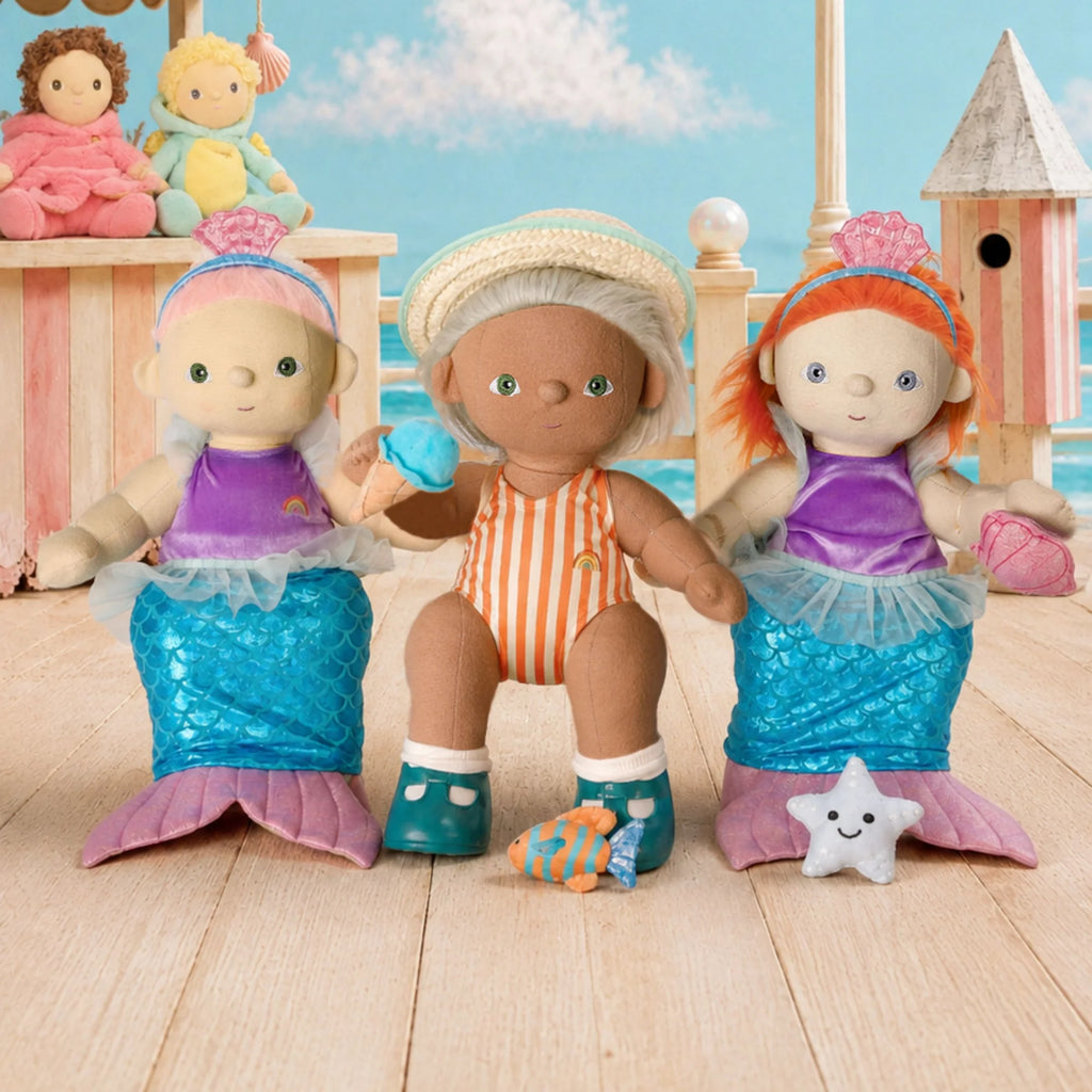 Three mermaid-themed baby dolls seated on a wooden deck with a beach and house backdrop. The center doll wears an orange-and-white striped swimsuit and cream hat, while the two side dolls wear purple tops and blue mermaid tails with pink fin accents.