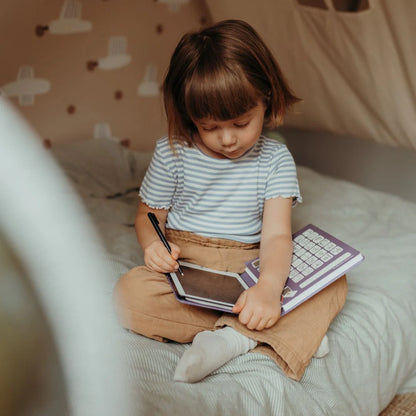 Child sitting on a bed with notebooks and a pen, in a cozy room setting.