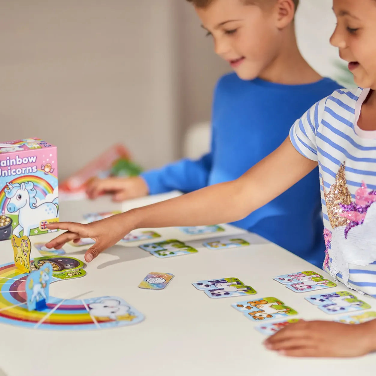 Two children playing a board game with colorful cards and a box labeled 'Rainbow Unicorns' on a table.