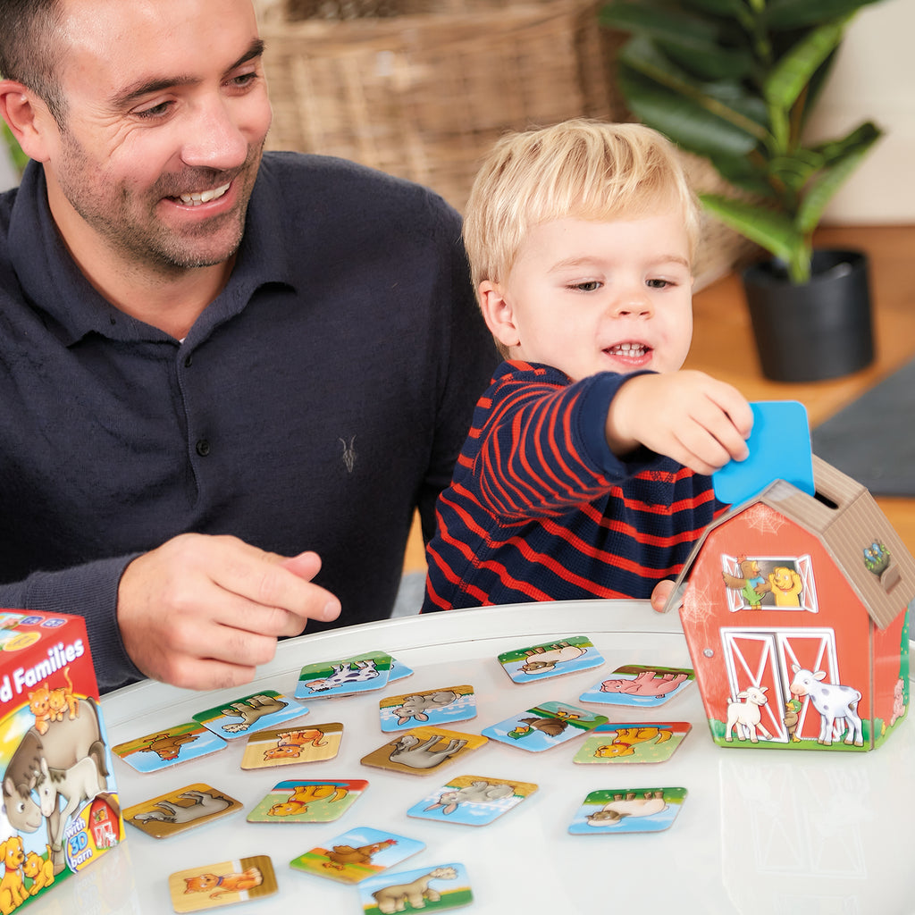 Man and child playing with a toy barn and card game on a table.