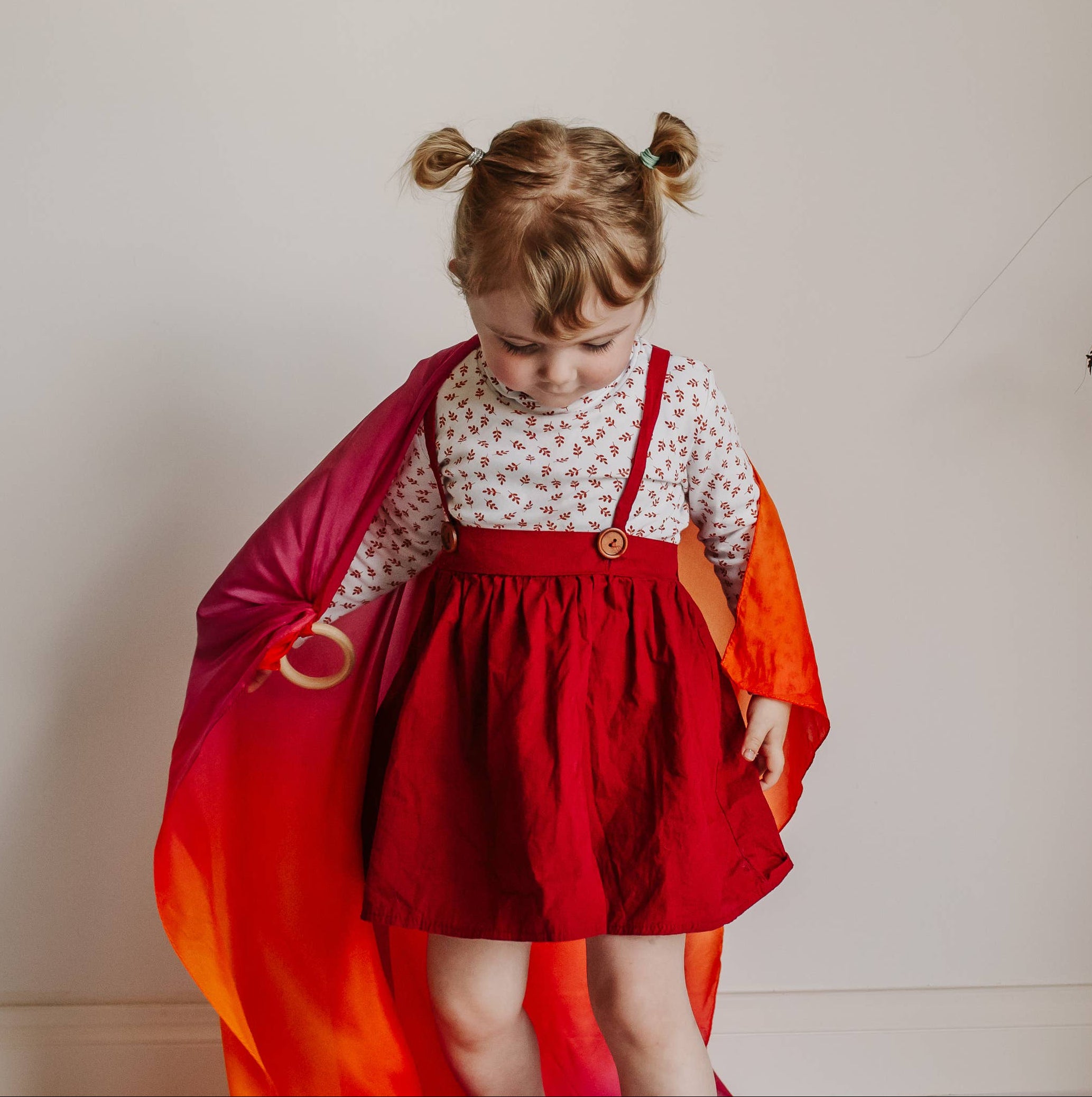 Child in a red dress with a colorful cape standing next to a decorative plant against a white wall.