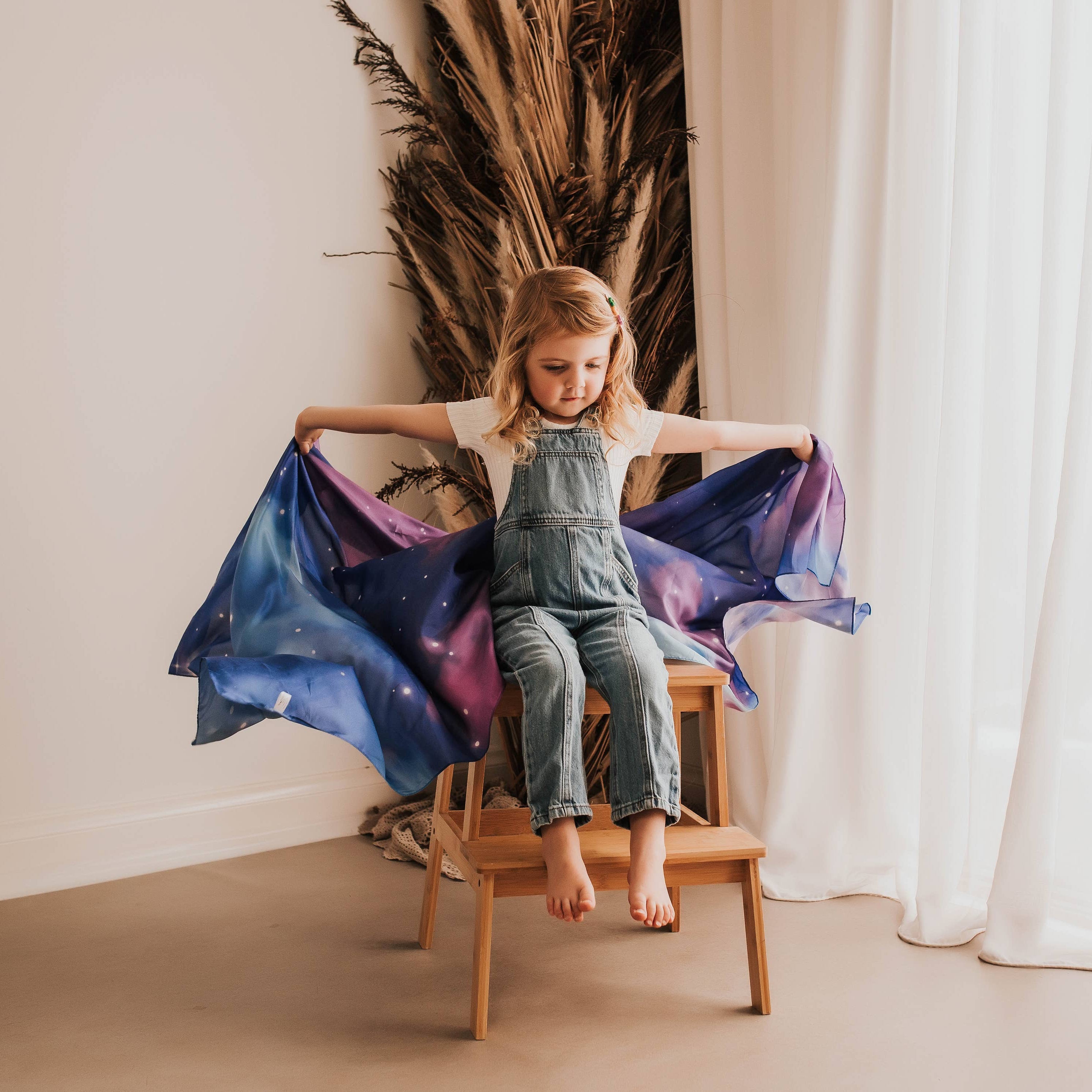 A young girl with blonde hair sits on a wooden stool in a bright room, holding up a large matching scarf or fabric piece with galaxy print in blue, purple, and pink. She is wearing denim overalls and a white t-shirt.