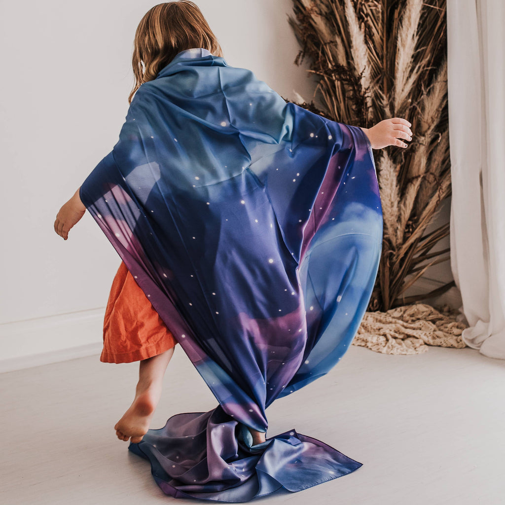 A child seen from behind wearing a vibrant galaxy-printed wrap or shawl with blue, purple, and pink nebula patterns and white star details. The child is standing in a minimalist room with a white wall, light floor, and a dried palm plant in the background.