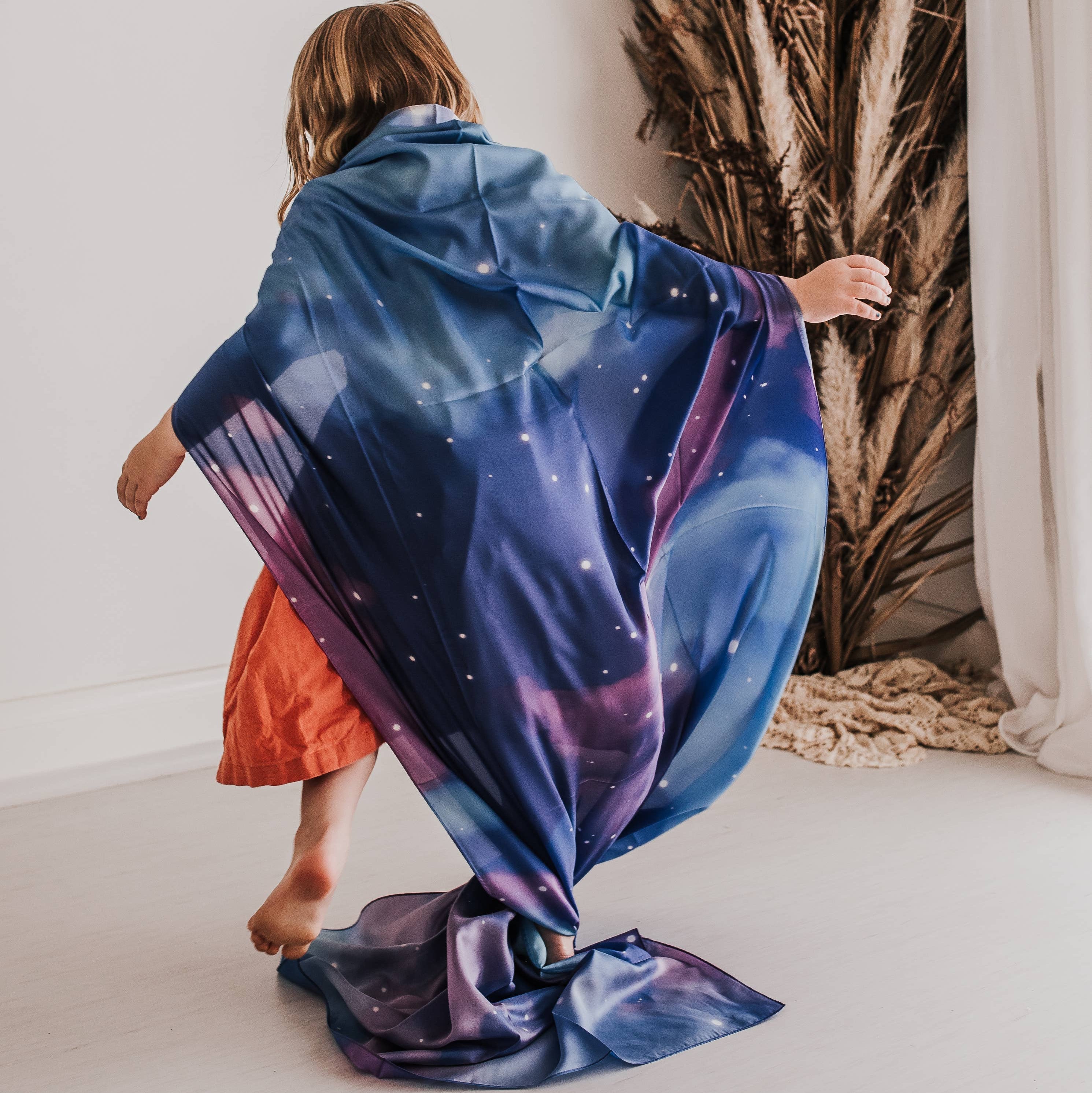 A child seen from behind wearing a vibrant galaxy-printed wrap or shawl with blue, purple, and pink nebula patterns and white star details. The child is standing in a minimalist room with a white wall, light floor, and a dried palm plant in the background.