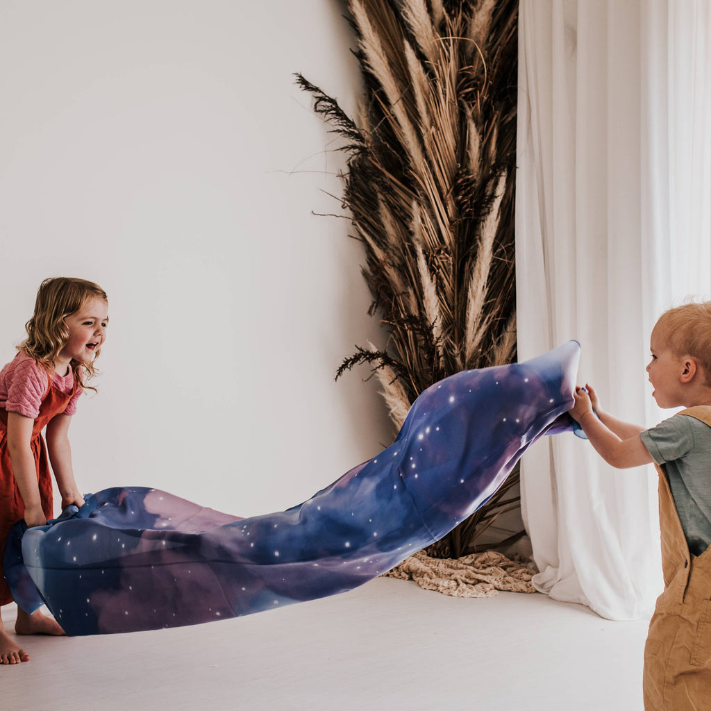 Two children playing with a long, lightweight fabric blanket printed with a galaxy pattern in shades of blue, purple, and white, set in a bright, modern room with white walls and curtains.