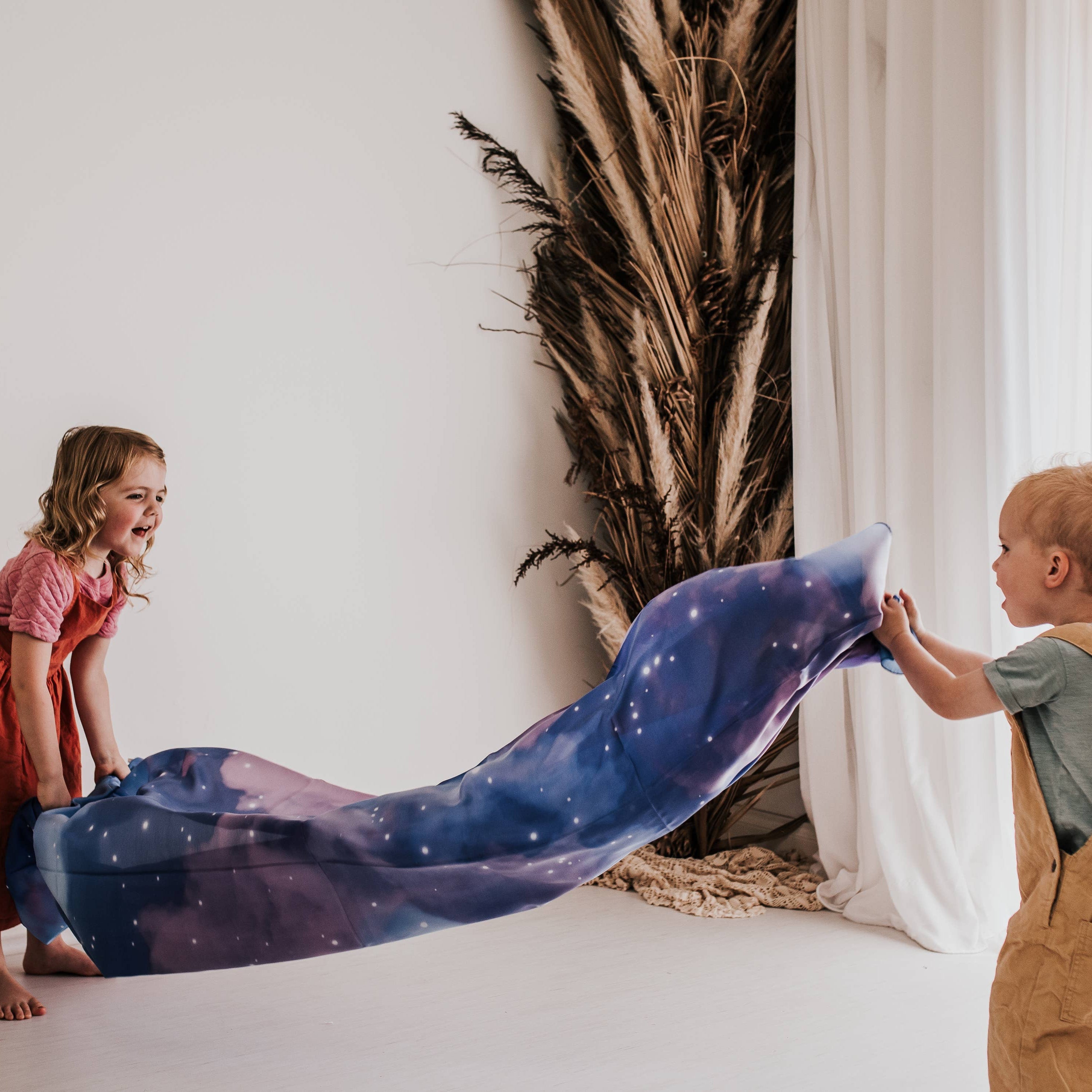 Two children playing with a long, lightweight fabric blanket printed with a galaxy pattern in shades of blue, purple, and white, set in a bright, modern room with white walls and curtains.