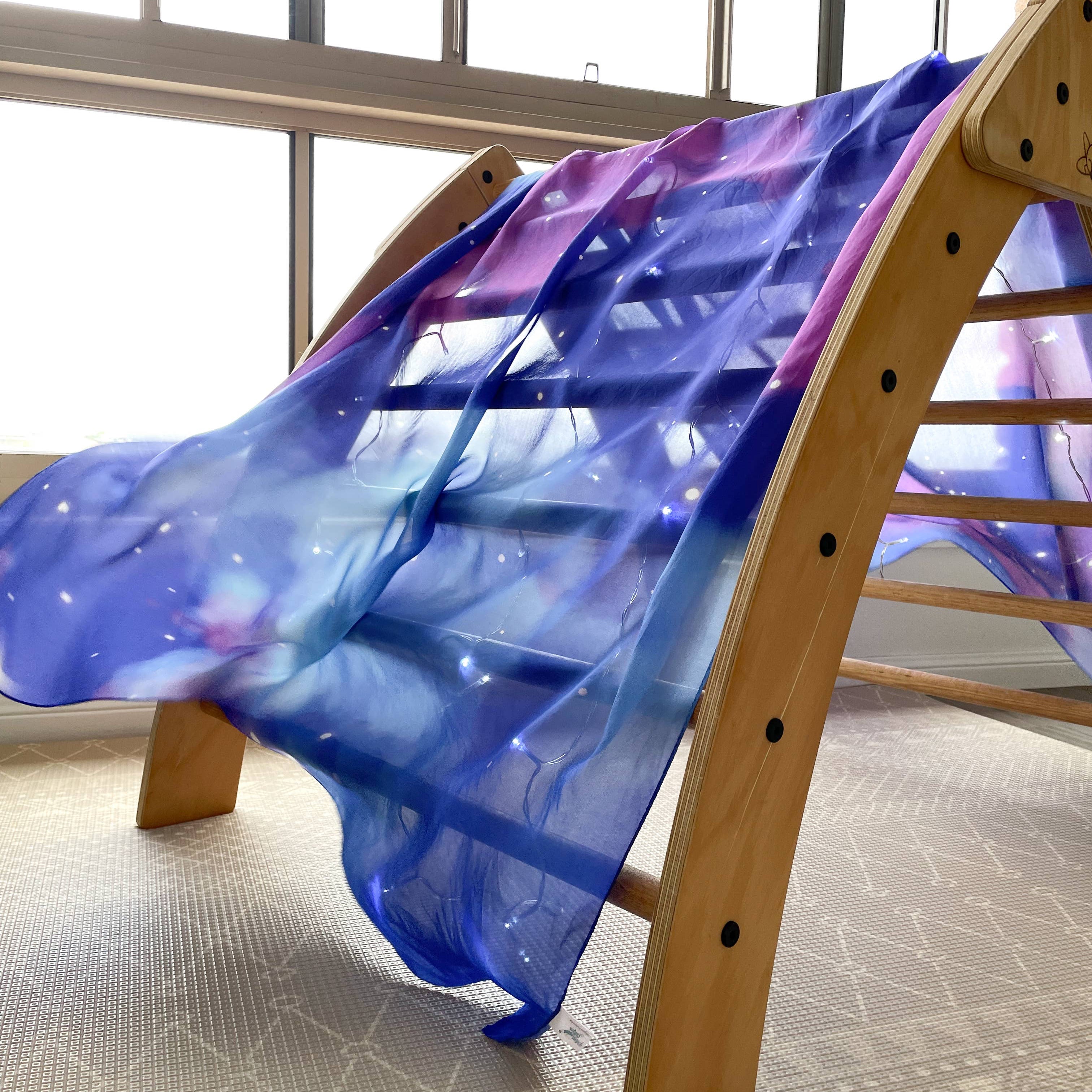 A sheer, flowing fabric with a galaxy print in shades of blue, purple, and white, draped over the side of a wooden climbing frame in a bright indoor setting.