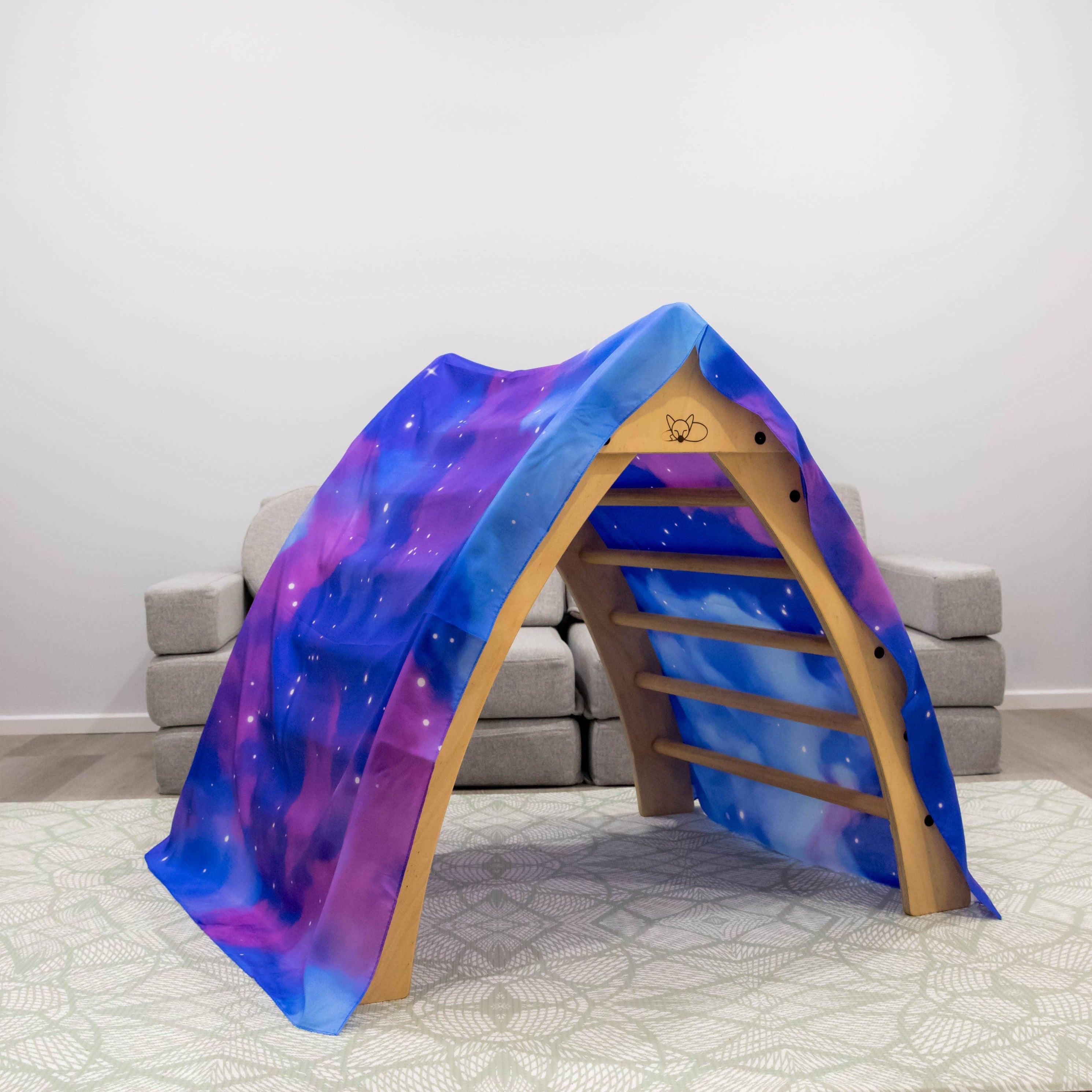 A wooden play tunnel with a colorful galaxy-print fabric roof and side panels, set on a light rug in a minimalist room with gray block seating in the background.