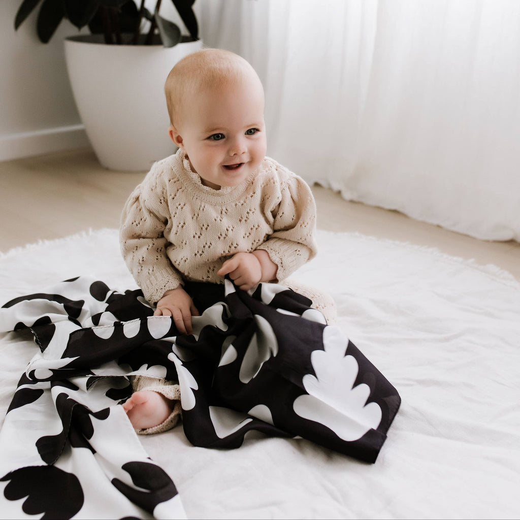 Baby sitting on a black and white patterned blanket with a plant in the background