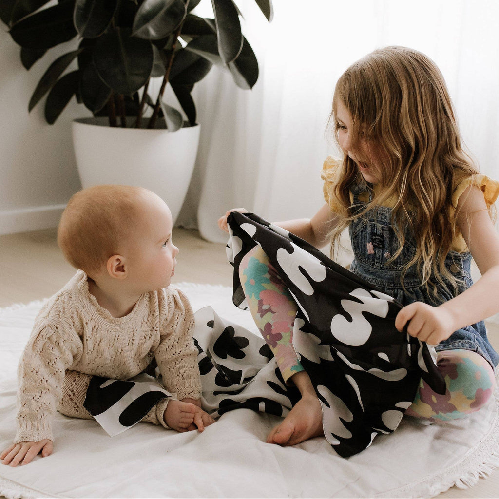 Two children playing with a black and white patterned blanket on a light-colored floor.