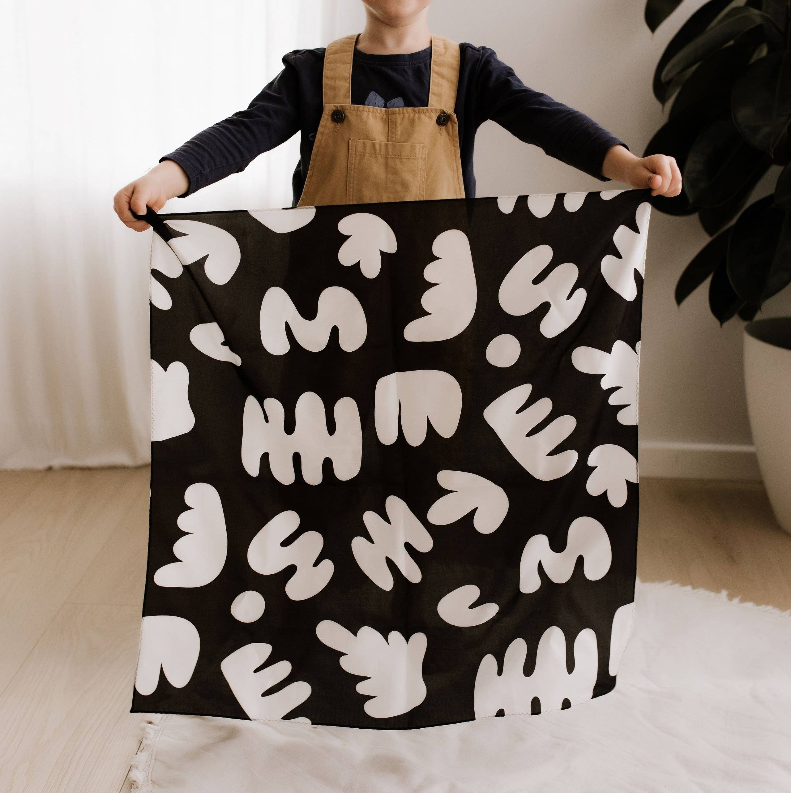 Child holding a black blanket with white abstract patterns in a room with a plant and rug.