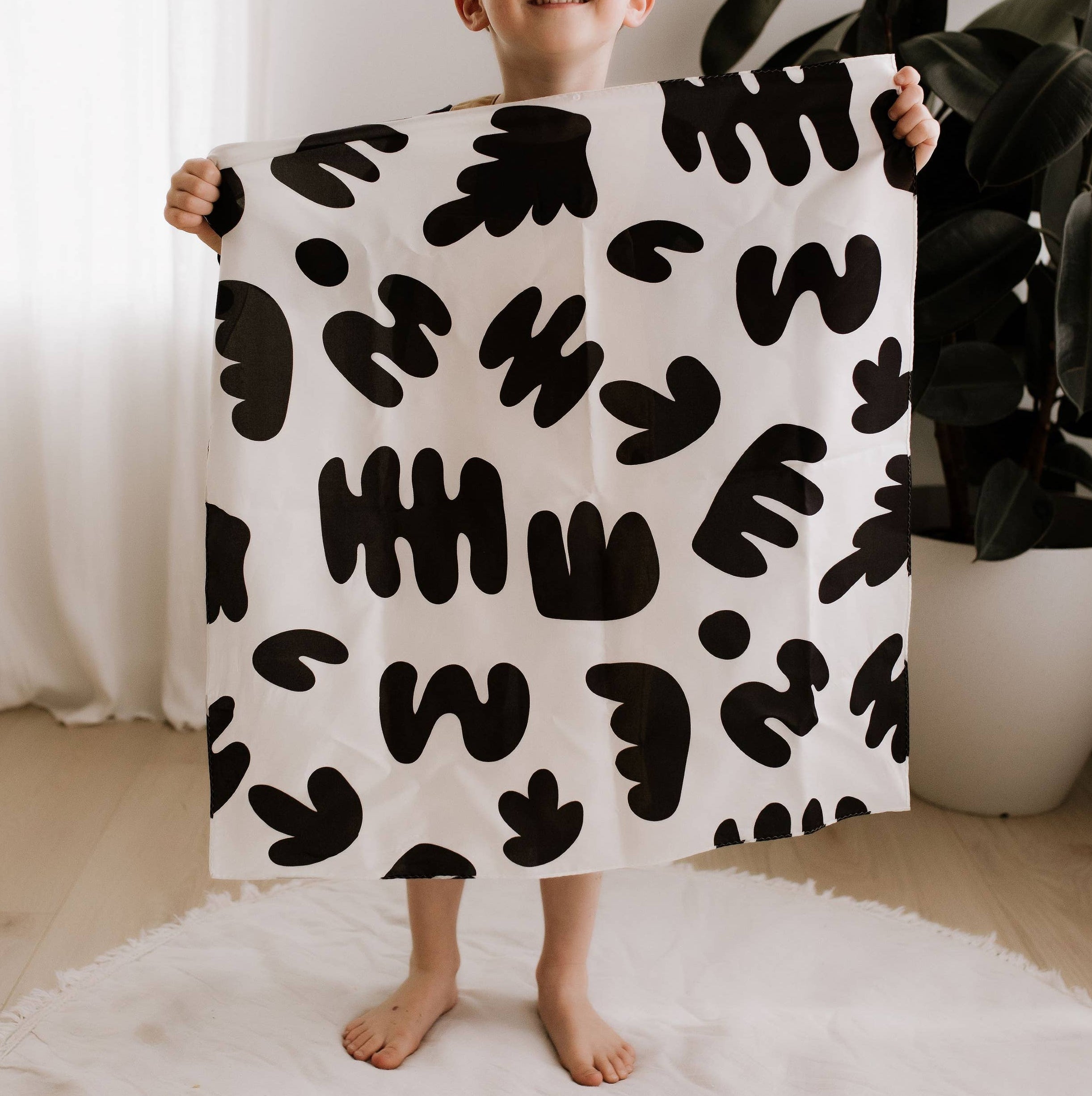 Child holding a black and white patterned blanket in a room with a plant and white curtain.