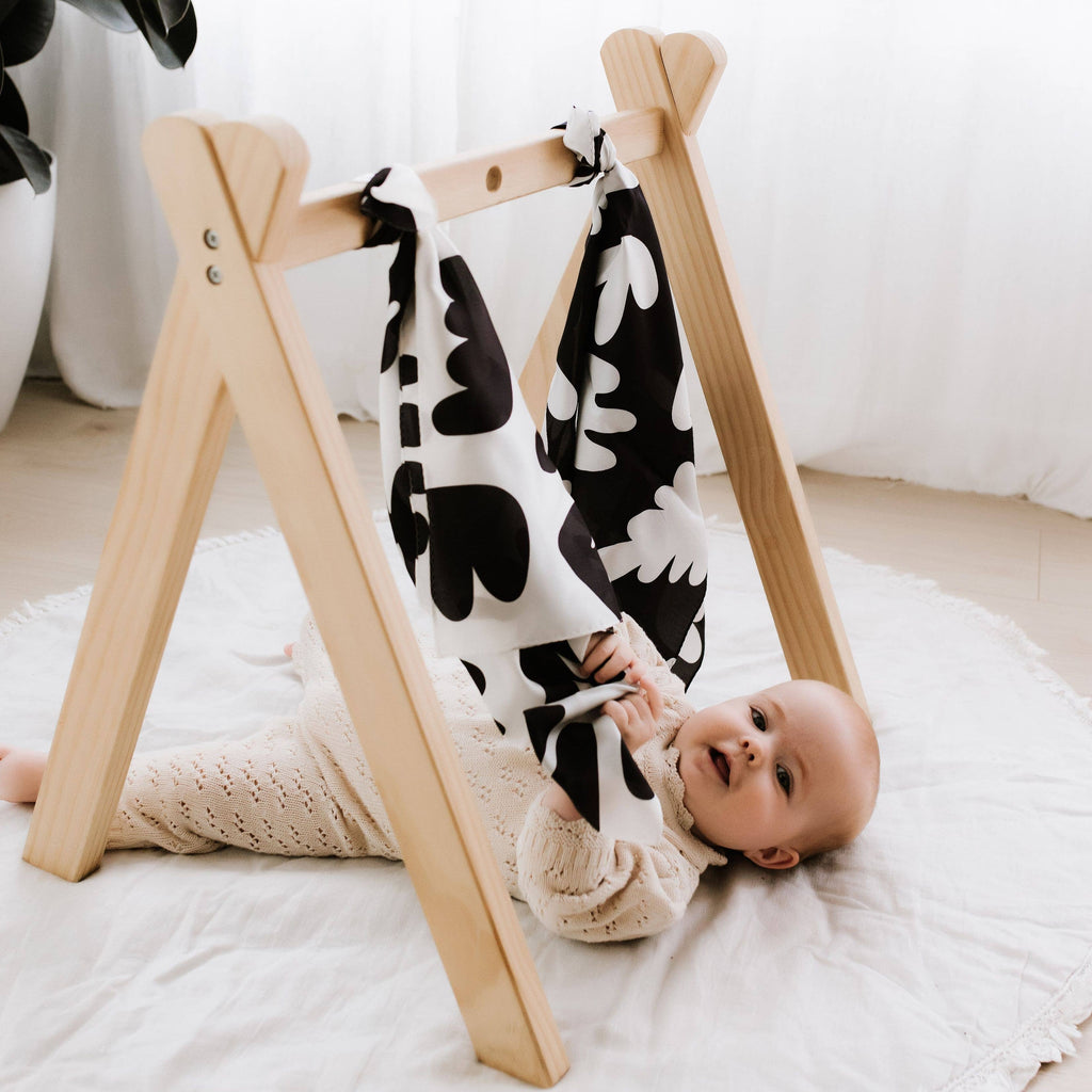 Baby lying on a white blanket next to a wooden baby gym with a black and white patterned blanket draped over it.