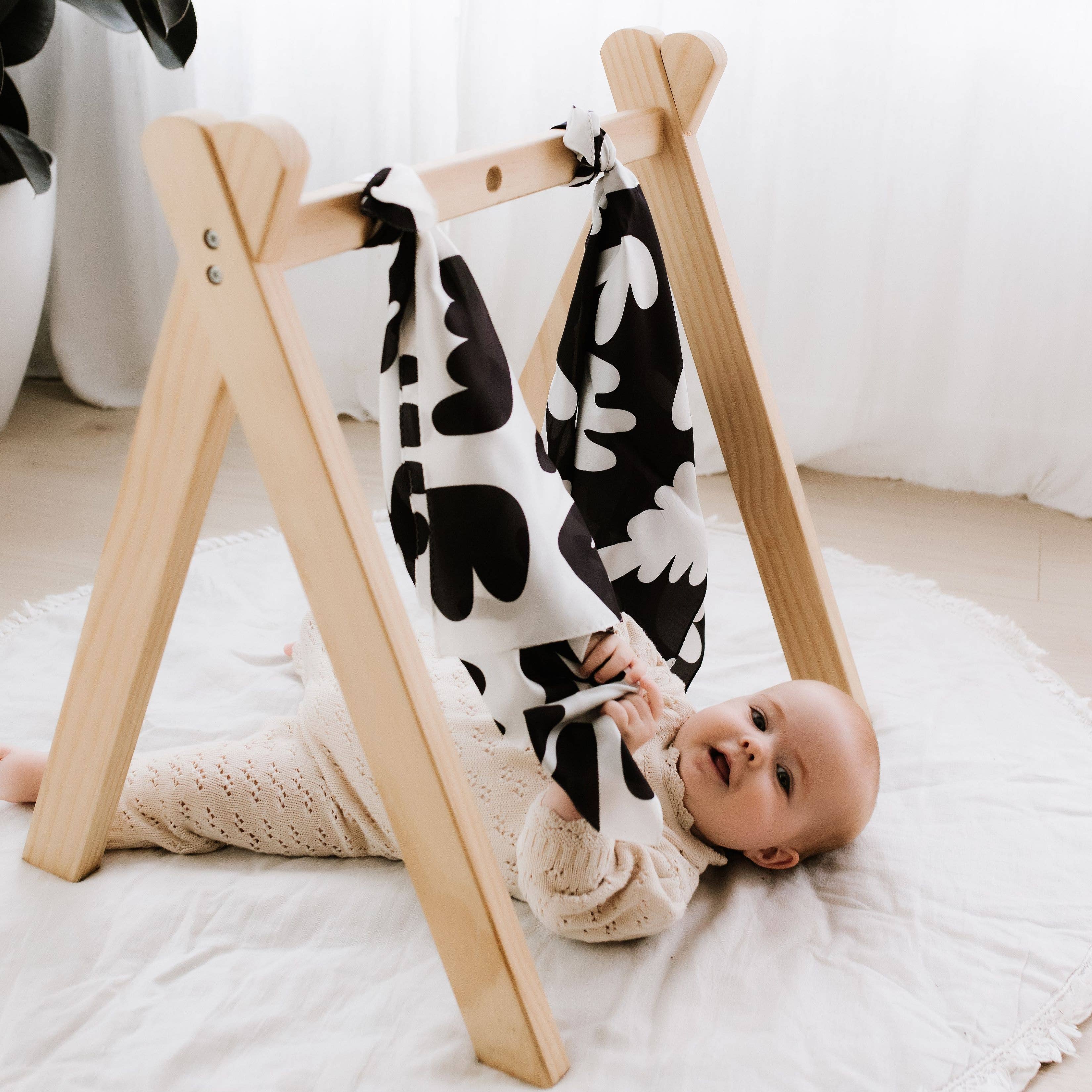 Baby lying on a white blanket next to a wooden baby gym with a black and white patterned blanket draped over it.