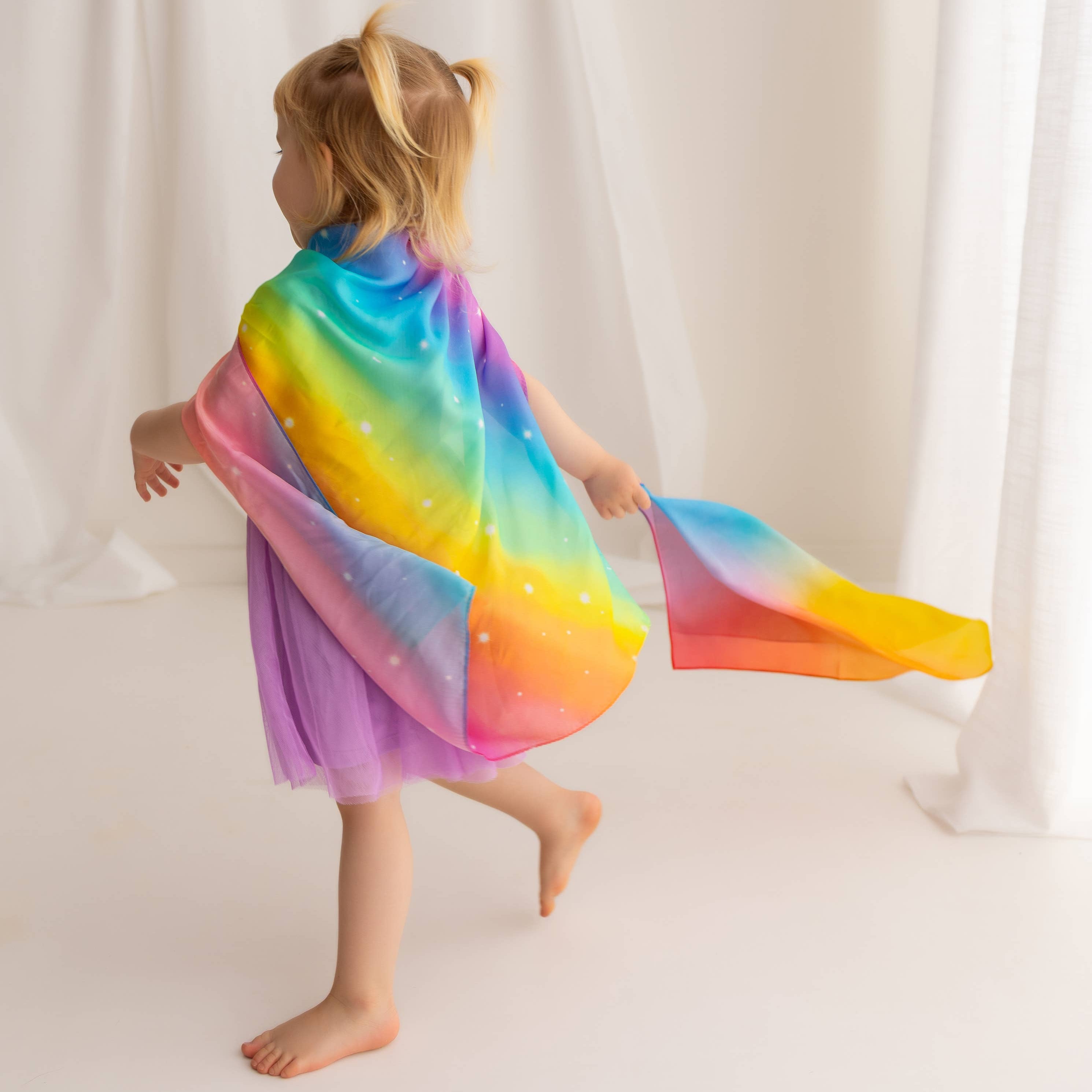 Child wearing a colorful rainbow cape against a white curtain background