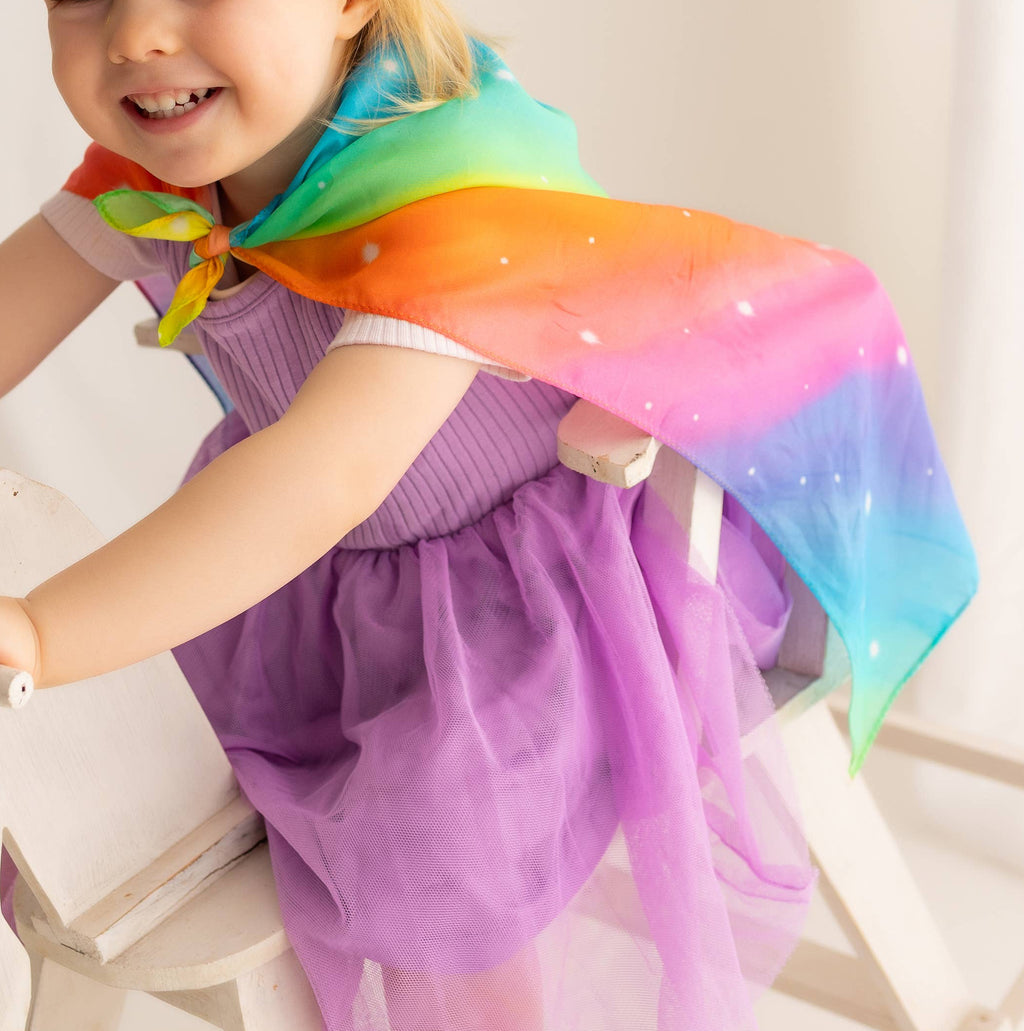 Child wearing a purple dress and rainbow scarf sitting on a white chair.