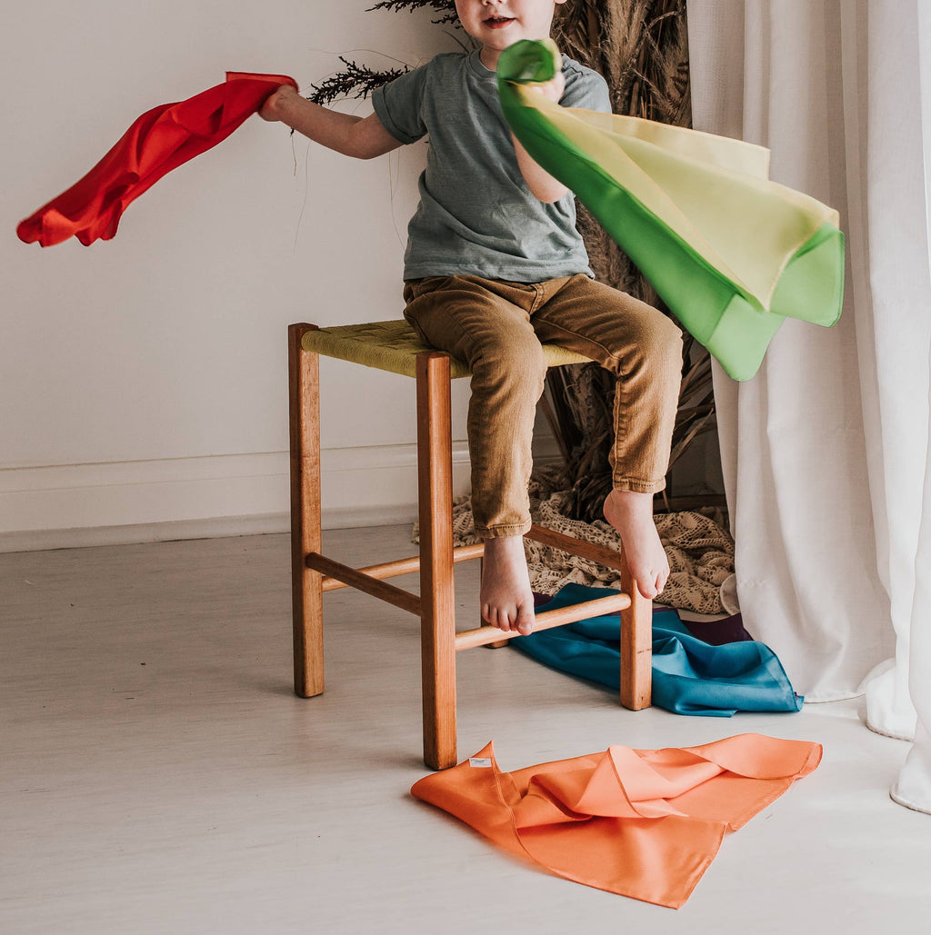 Child sitting on a stool holding colorful cloths in a room with decorative plants and curtains.