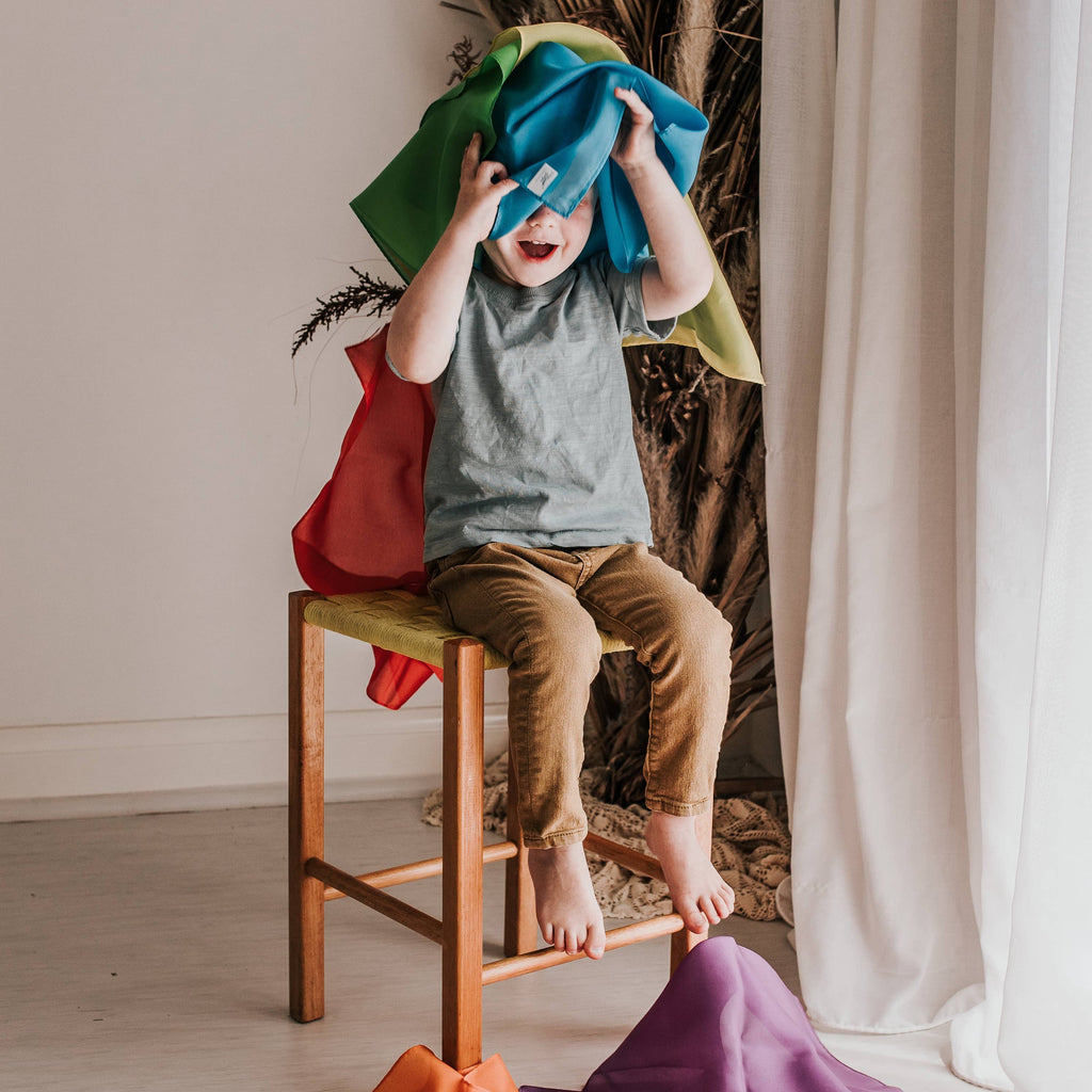 Child sitting on a stool with colorful cloths, surrounded by a decorative plant and draped fabrics.