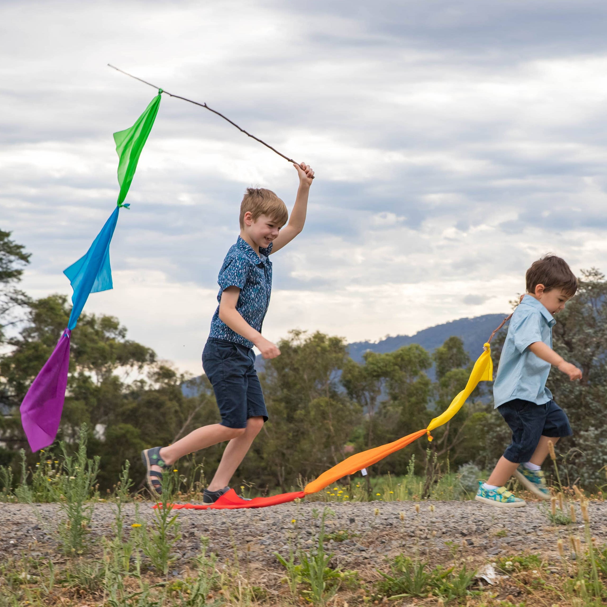 Two children flying a colorful kite in an open field with trees and mountains in the background.