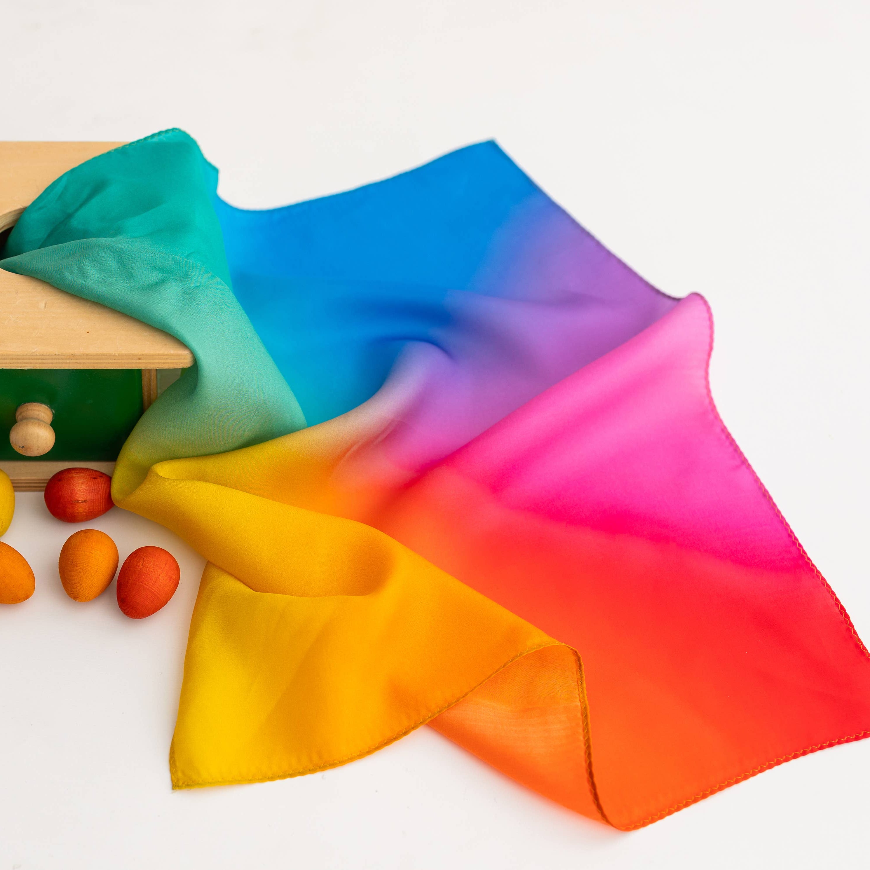 Colorful fabric flowing over a wooden toy with drawers and colorful balls on a white background