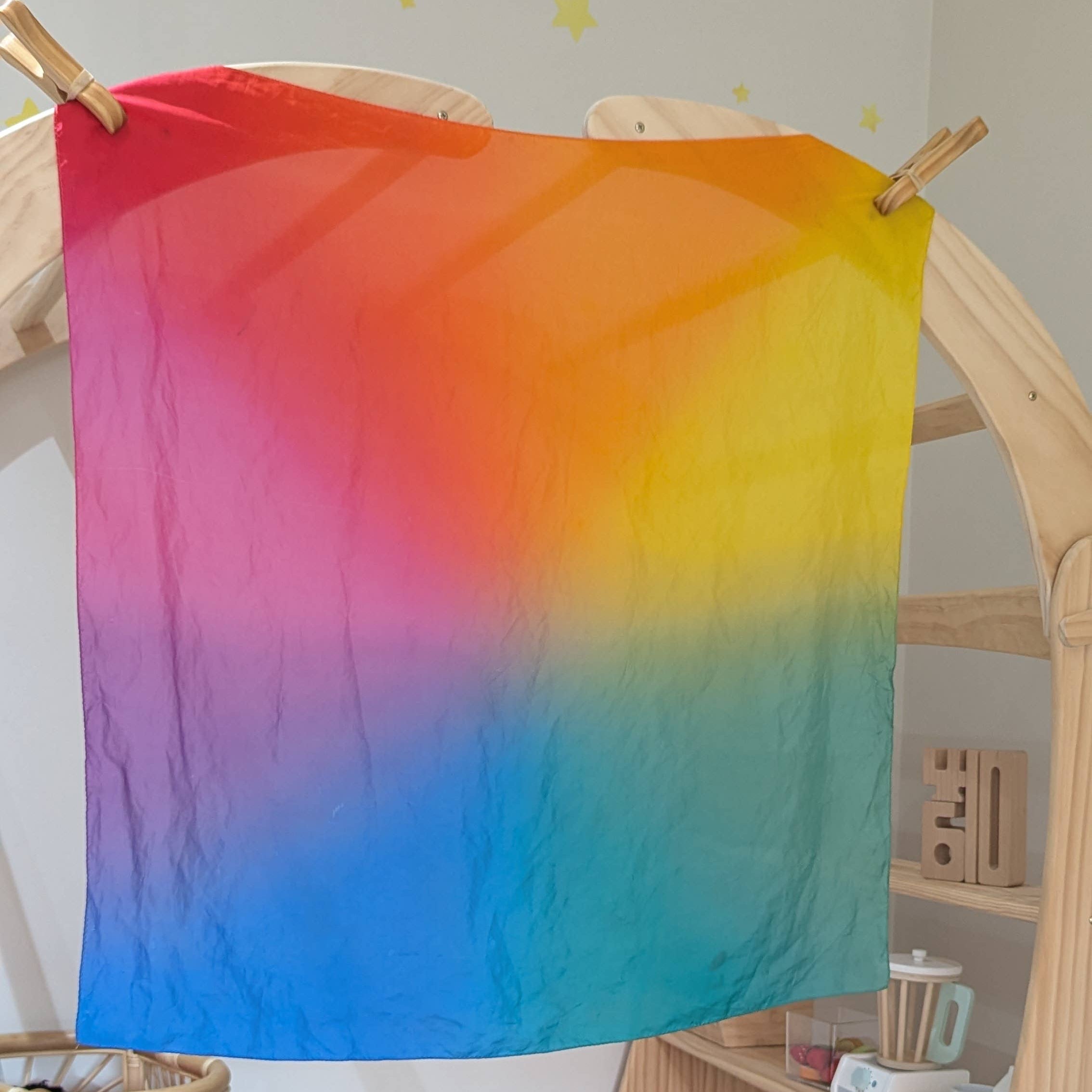 Colorful gradient fabric hanging on a wooden frame with toys and a shelf in the background.