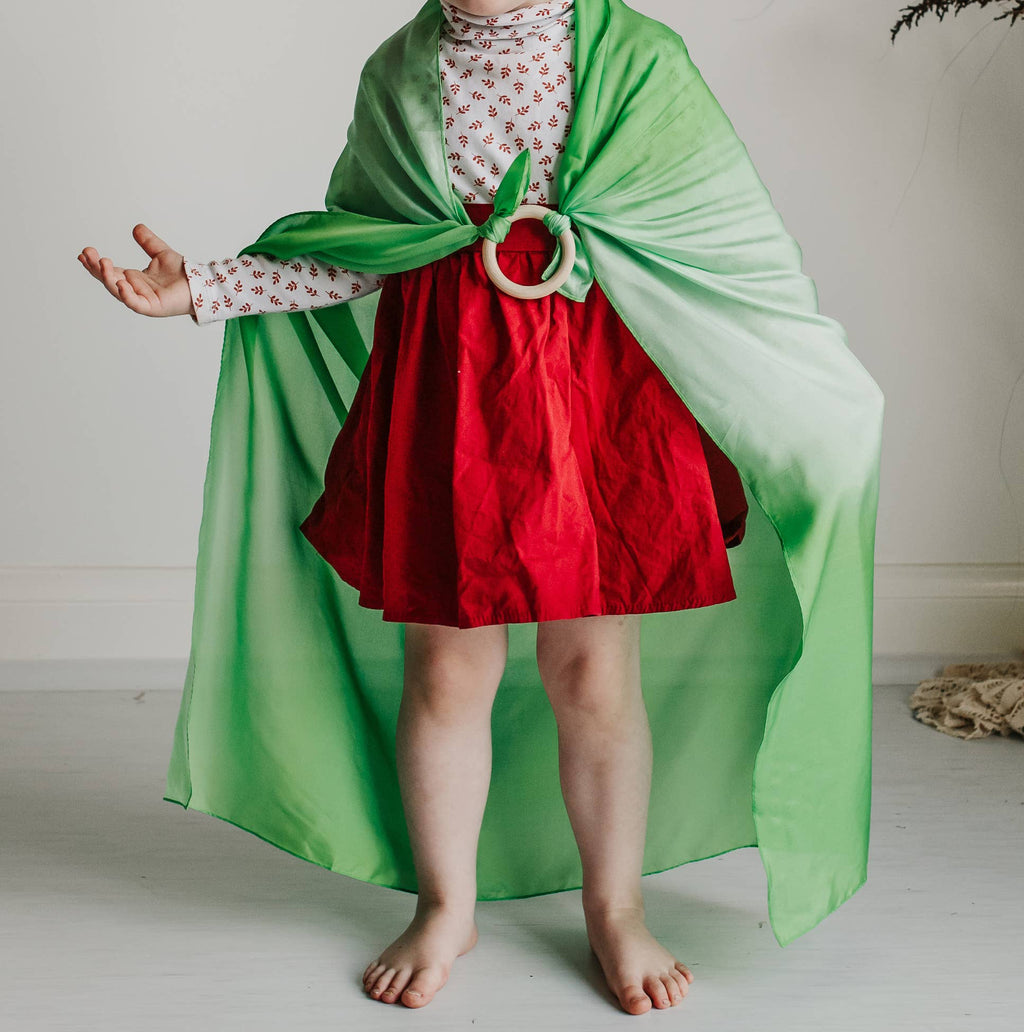 Child in a green cape and red dress standing in a room with a decorative plant.