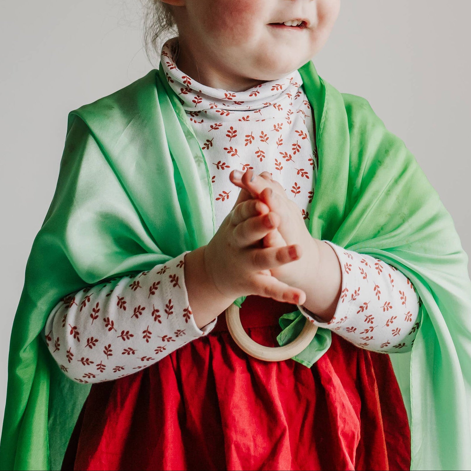 Young girl in a green shawl and red dress standing against a plain wall with a plant to the side.