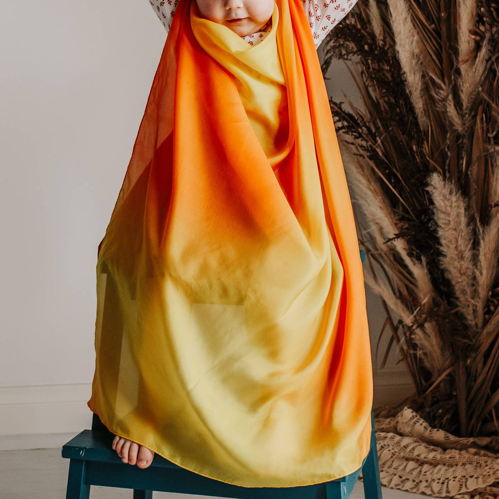 Child wrapped in a colorful blanket sitting on a stool with decorative plants in the background