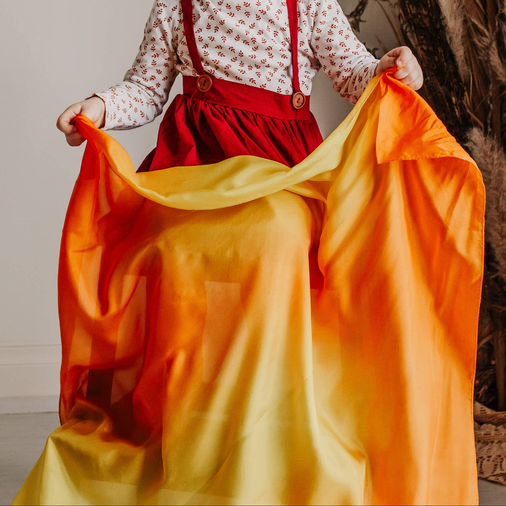 Child holding a colorful fabric in a room with a plant and white curtain.
