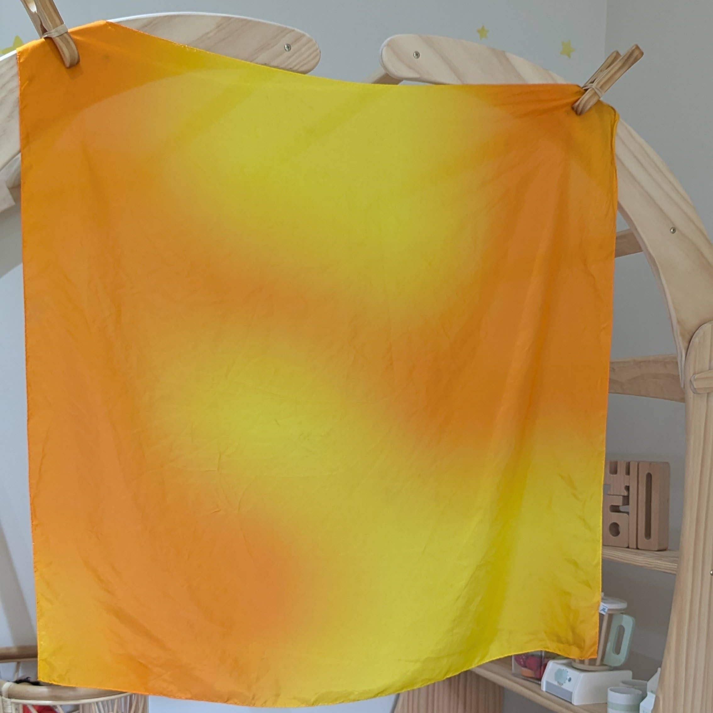 Yellow and orange gradient fabric hanging on a wooden rack with toys and a chair in the background.
